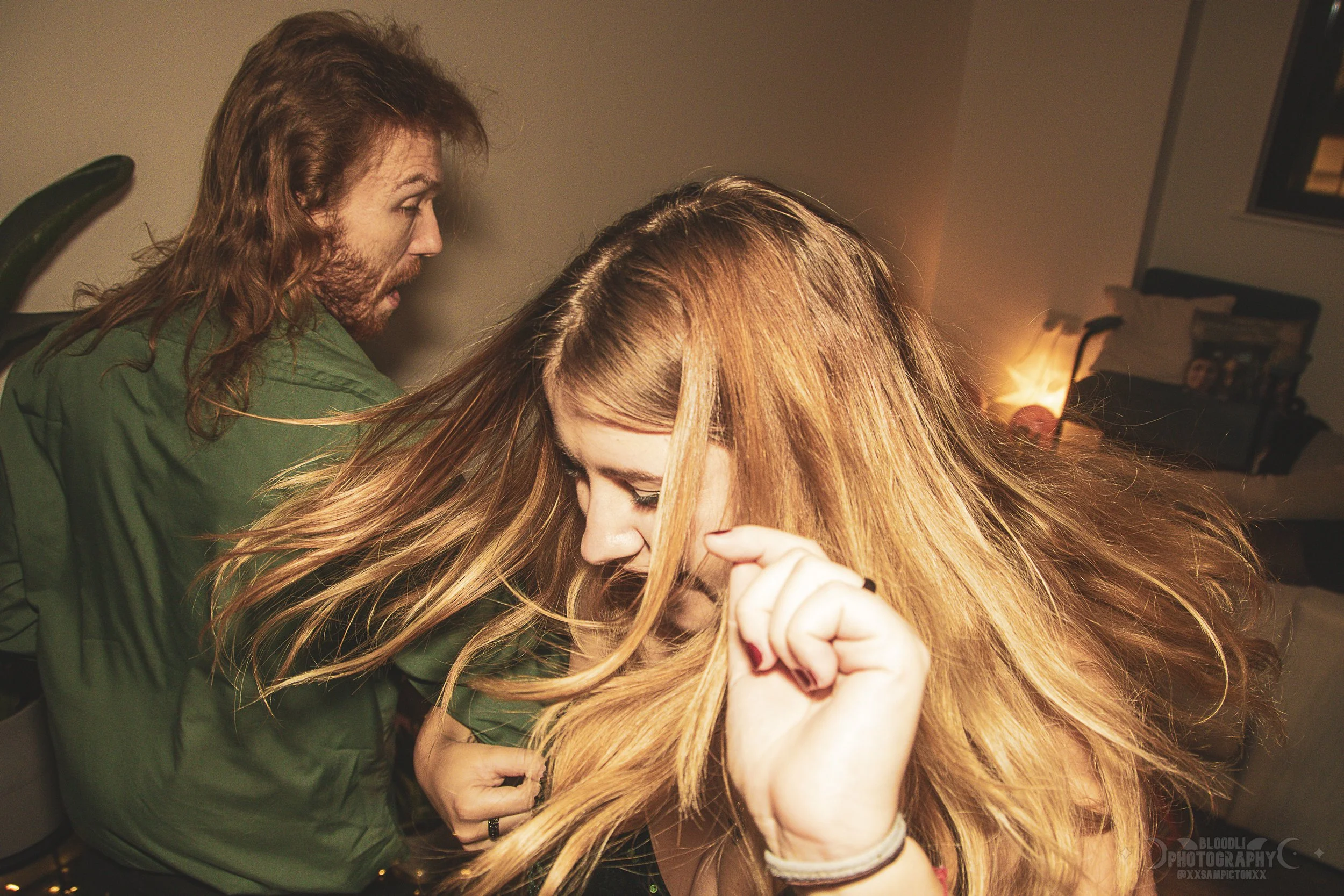 A young woman with long red hair dancing with her head tilted down, while a man with long brown hair and beard looking surprised on her left side in a room with warm lighting.