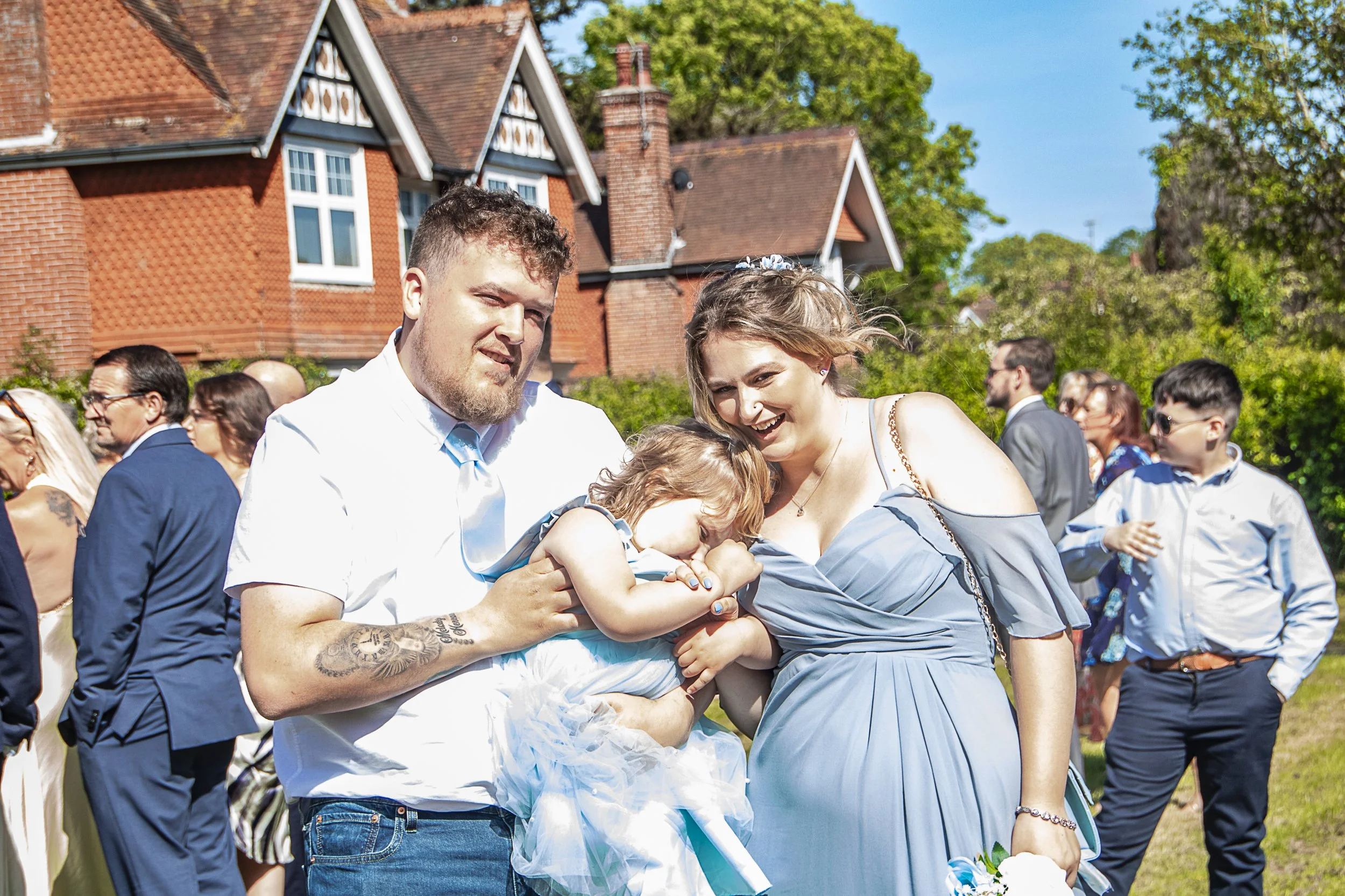 A group of people attending an outdoor wedding, with two adults holding and cuddling two children, surrounded by others dressed in formal attire on a sunny day with a red brick house and green trees in the background.