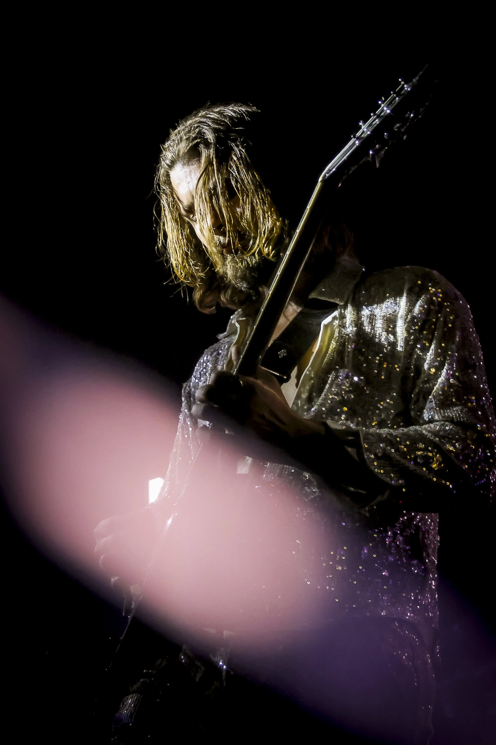 A musician with long blond hair playing an electric guitar on stage, wearing a shiny, sequined black jacket, under dark stage lighting.