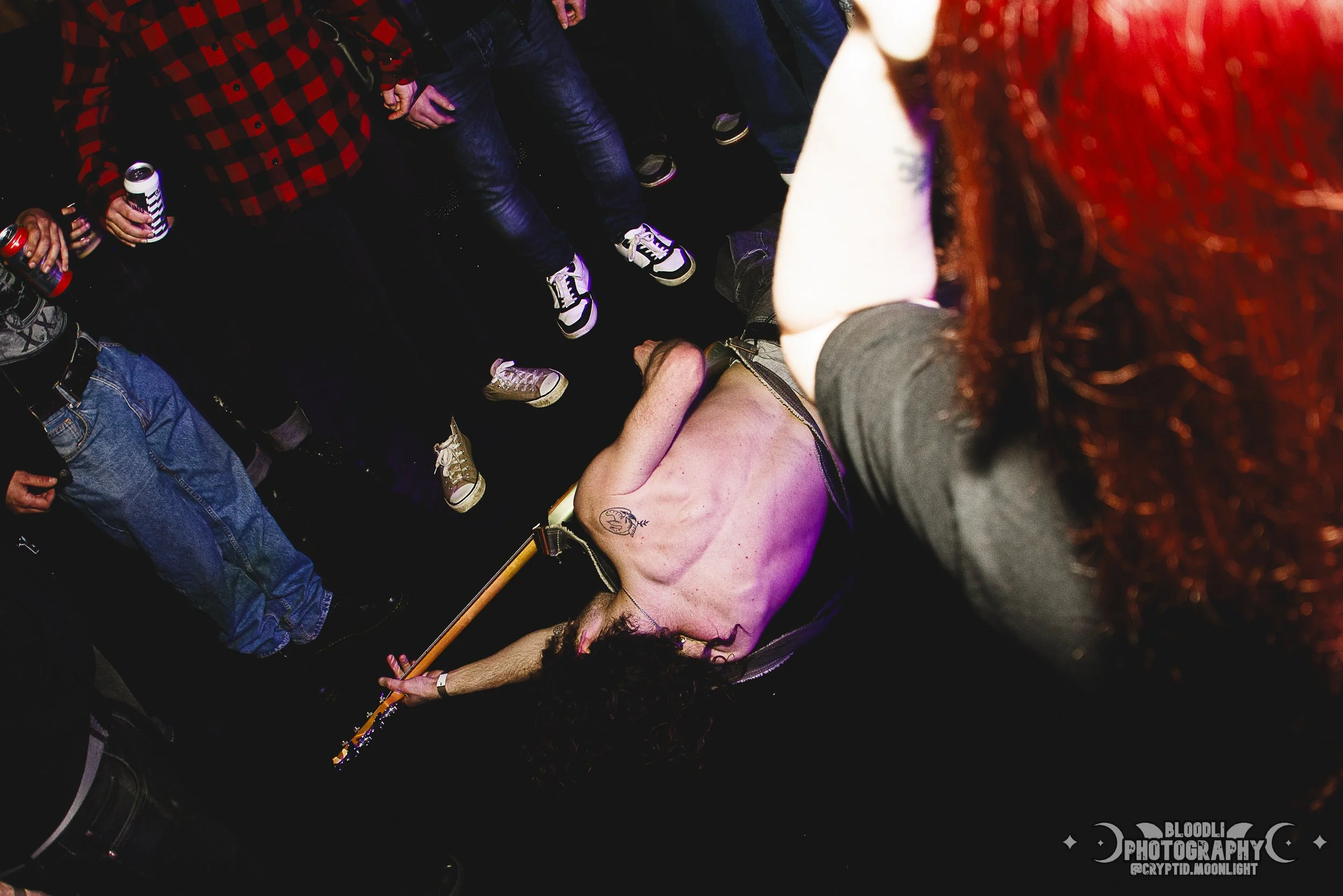 A shirtless person with long dark curly hair lying on the floor, surrounded by a crowd at a party or concert, holding a guitar.