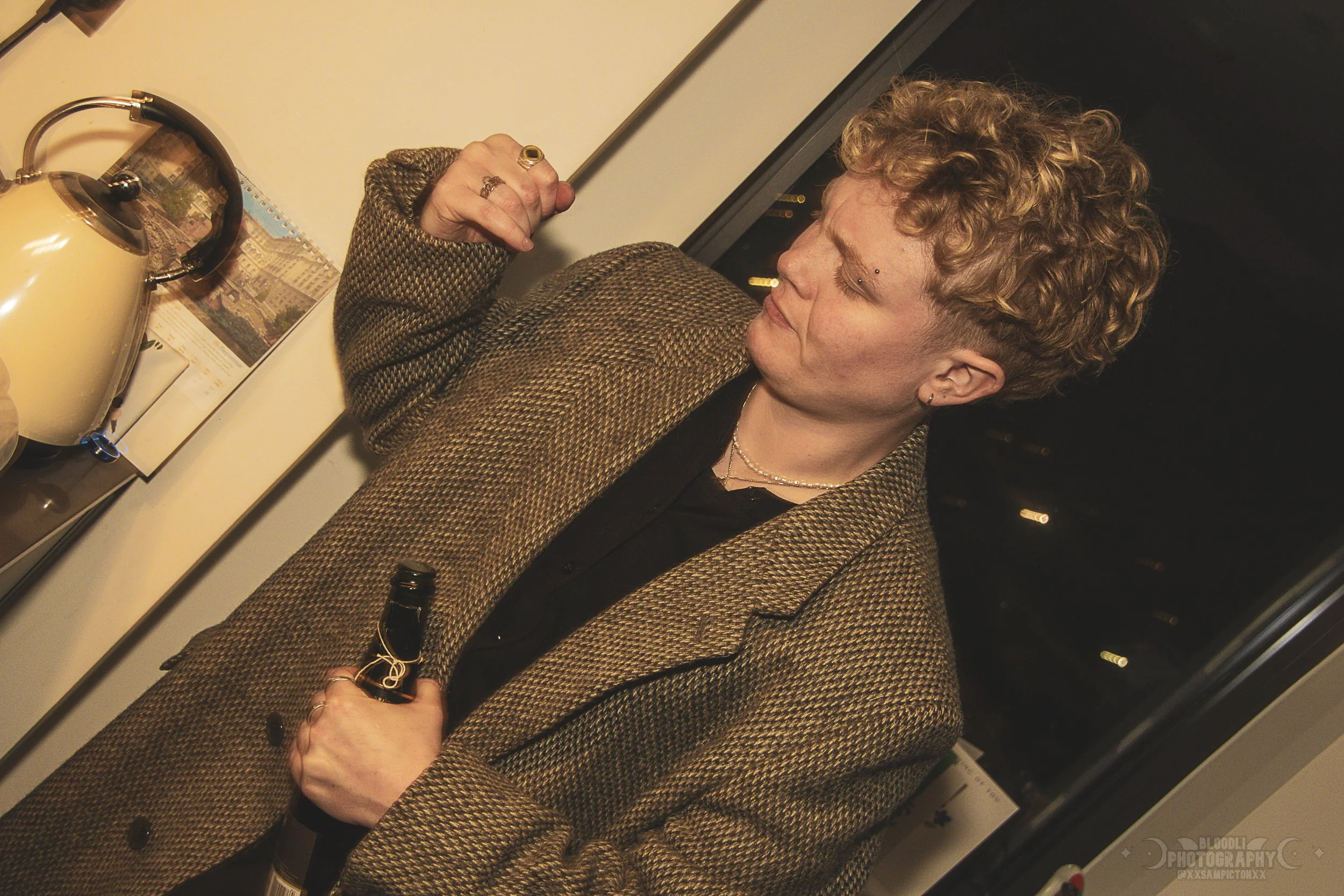 A person with curly blonde hair wearing a brown checkered blazer and black shirt, holding a beer bottle, standing at a bar with a cream-colored teapot and a small bottle on the bar counter, and a cityscape view through a window in the background.