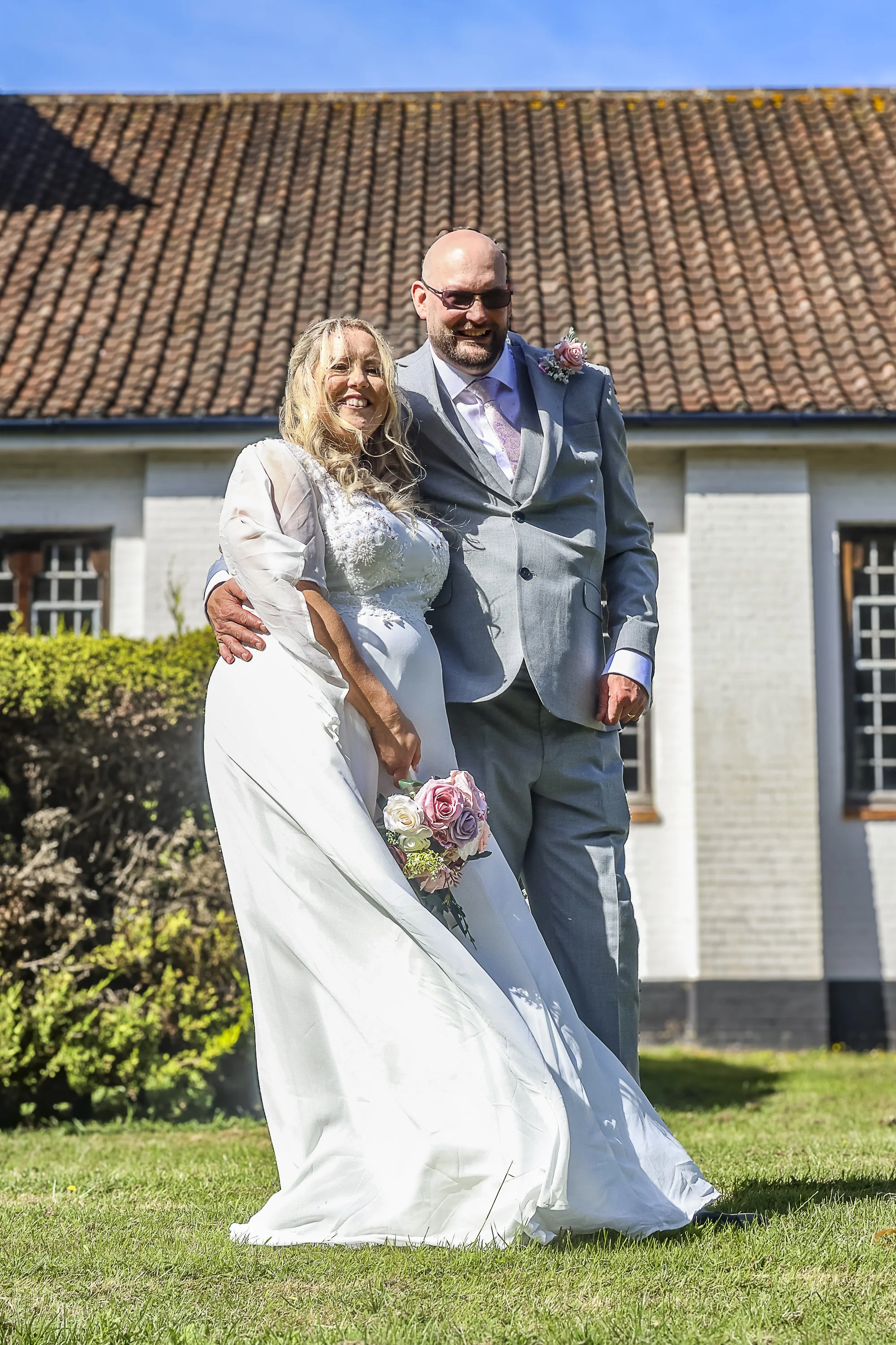 A smiling bride and a happy groom standing together outdoors on a sunny day in front of a house with a tiled roof, holding a bouquet of pink and white roses, with greenery and bushes in the background.