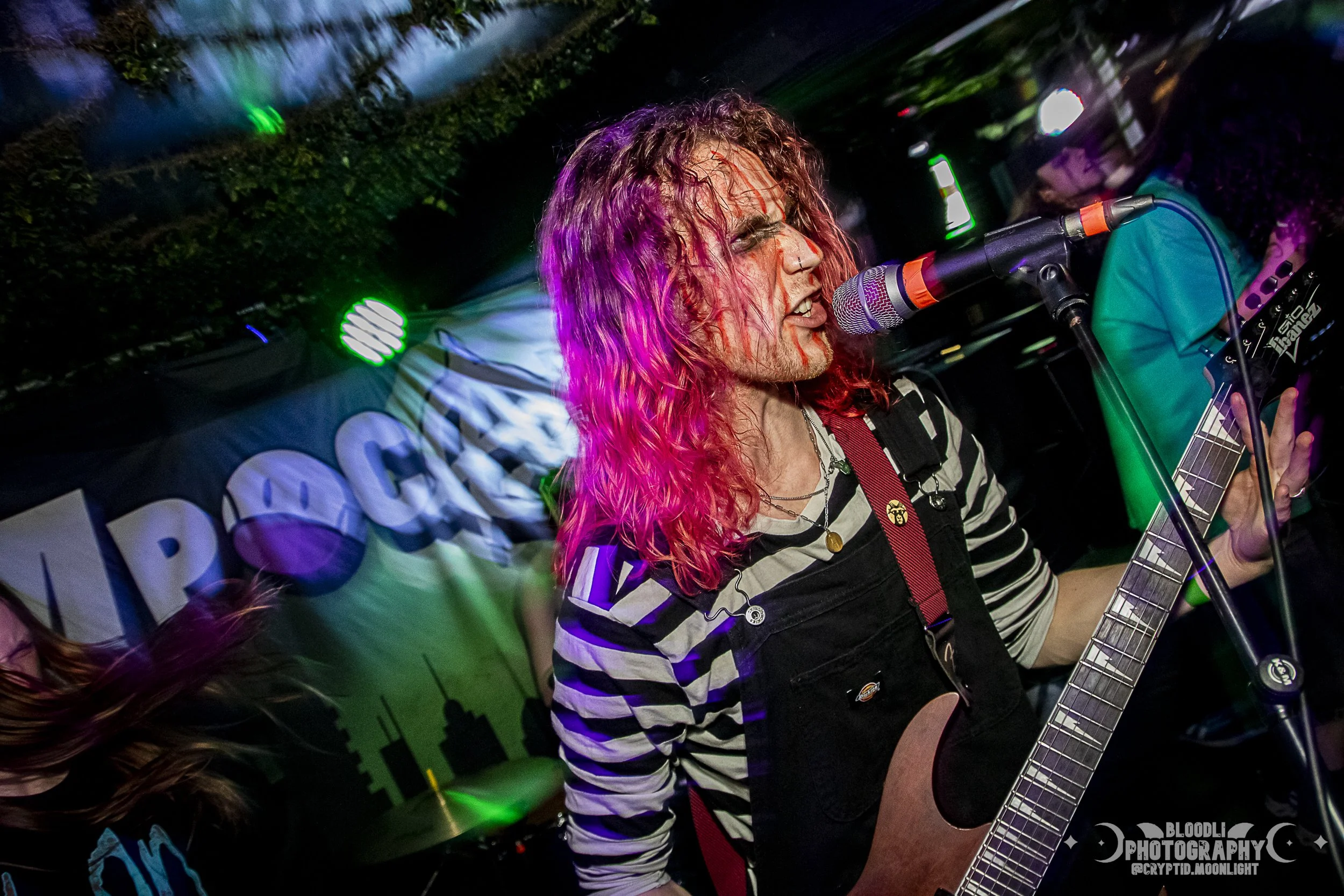 A musician with long, curly, pink hair passionately singing into a microphone while playing an electric guitar during a live performance. The background features colorful stage lighting and a banner with the word 'IMPOSSIBLE'.