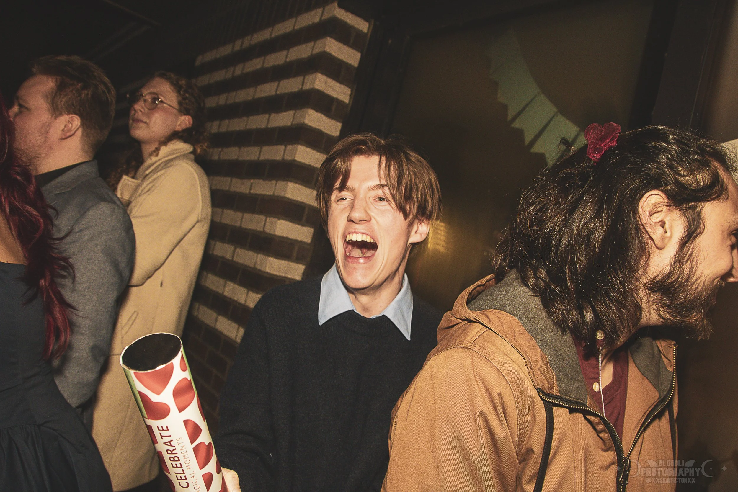 People at a celebration event, with a man laughing and holding a cylindrical paper decoration, alongside others standing in line, against a brick wall background.