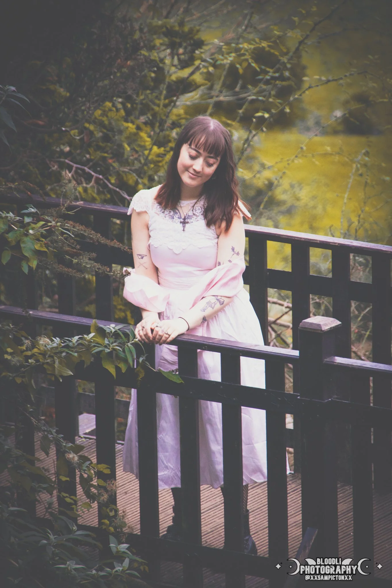 A young woman with tattoos and dark hair wearing a light pink dress, standing on a wooden bridge with black railings surrounded by greenery.