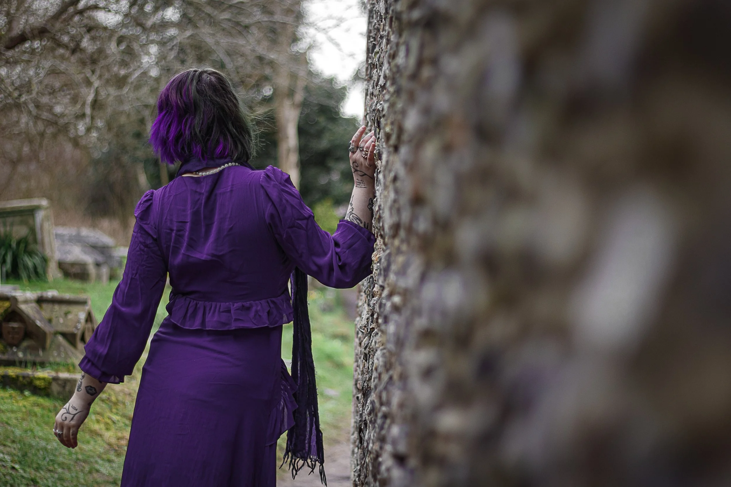 Person wearing a purple dress with tattoos on their arm, standing outdoors with their hand on a stone wall, facing away from the camera.