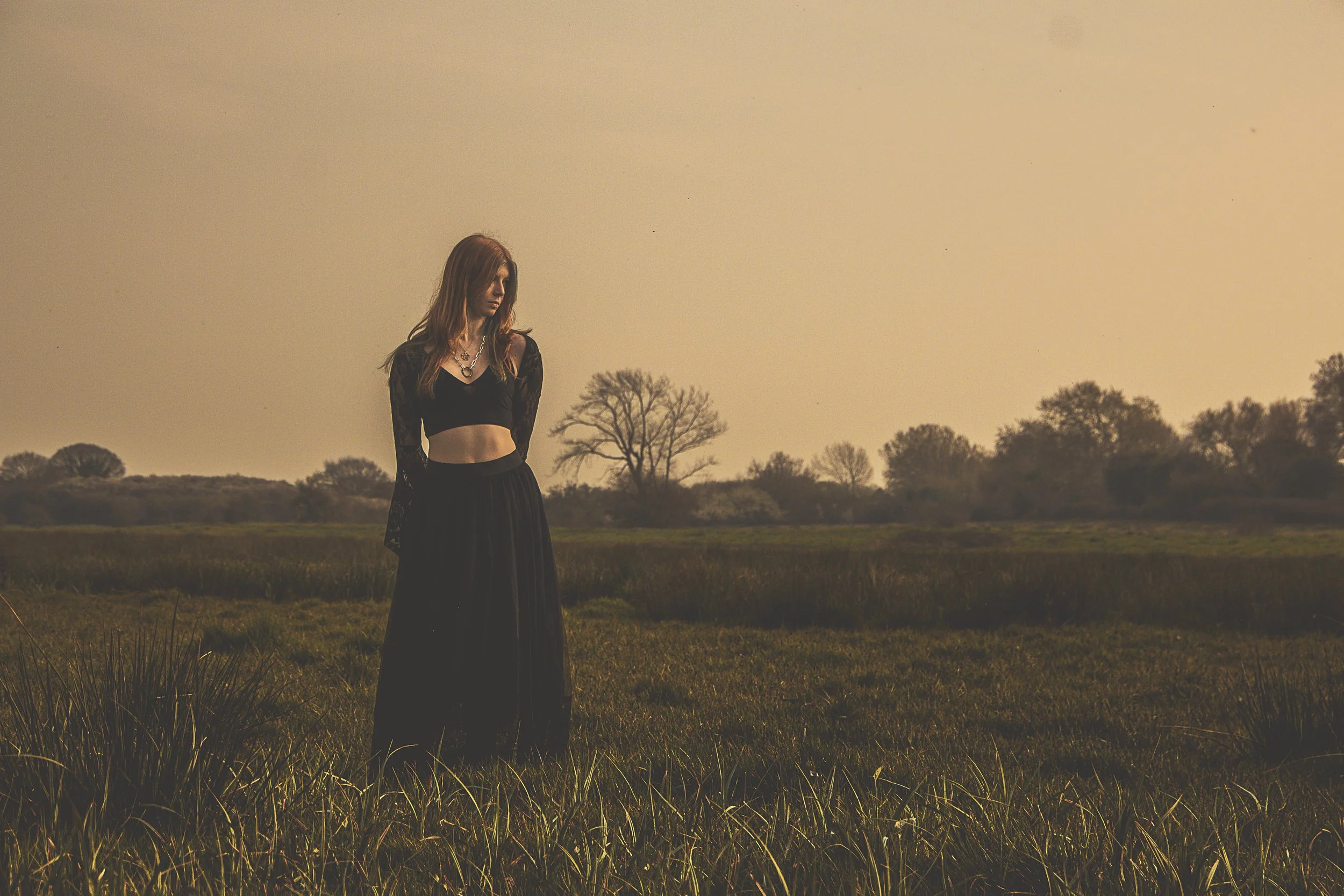 A young woman standing alone in a grassy field during sunset, wearing a black crop top with long lace sleeves and a long black skirt, with trees in the background.