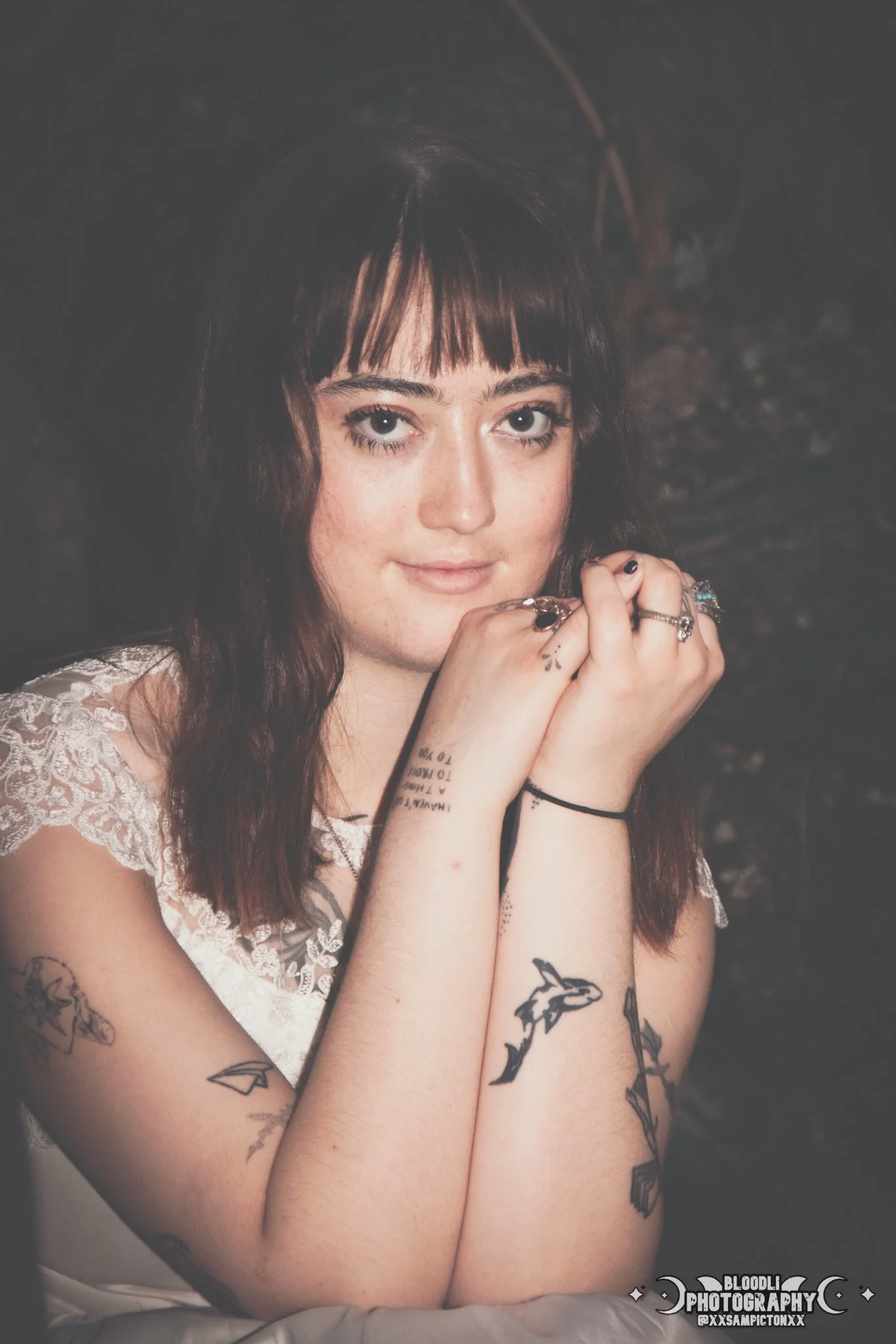 A young woman with dark brown hair and bangs, wearing a lace sleeveless top, resting her chin on her hands and smiling slightly. She has multiple tattoos on her arms including a dolphin, flowers, and geometric designs. The background is dark and blur