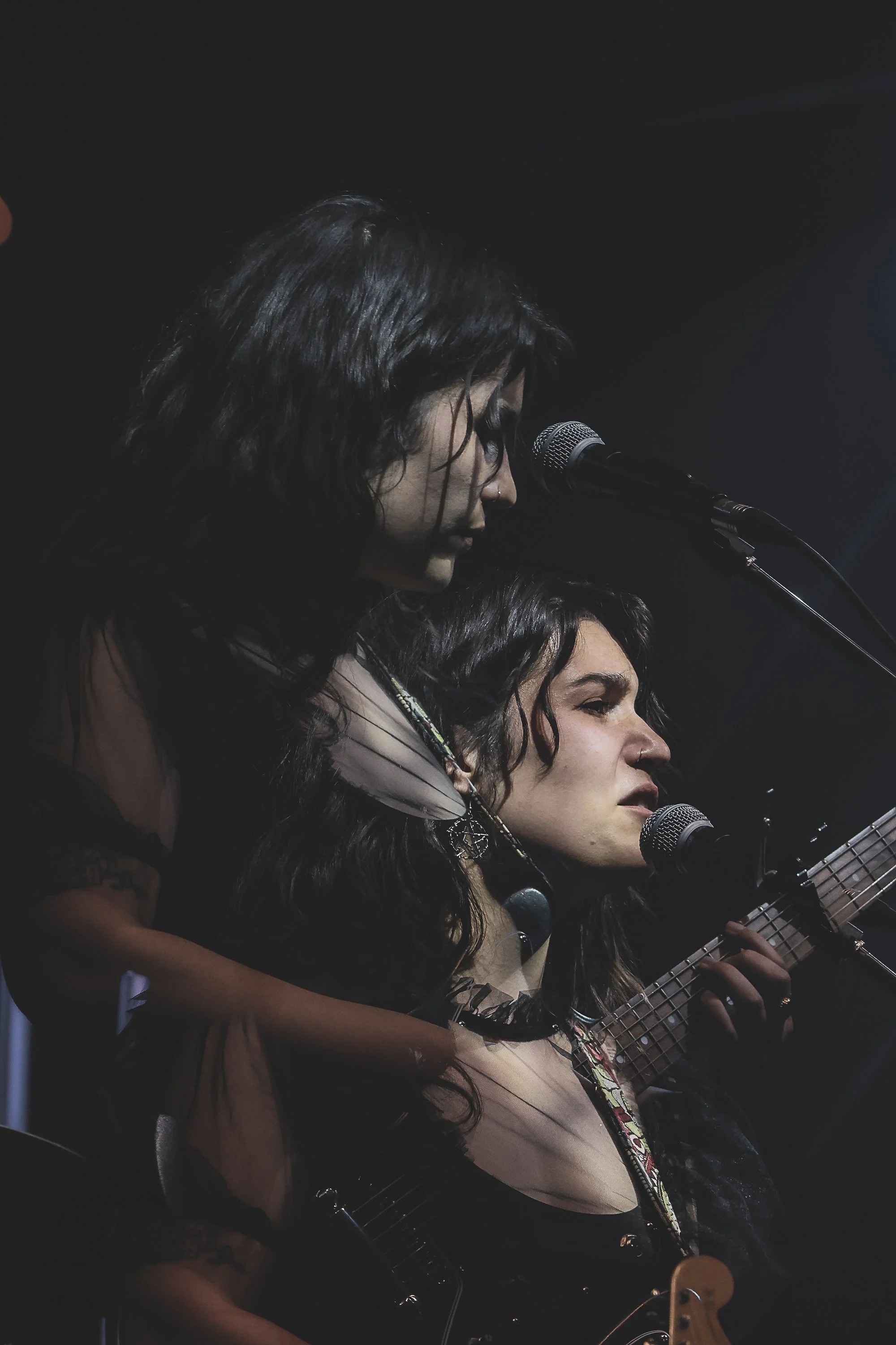 Two women singing into microphones, one playing an acoustic guitar, on stage in a dark background.