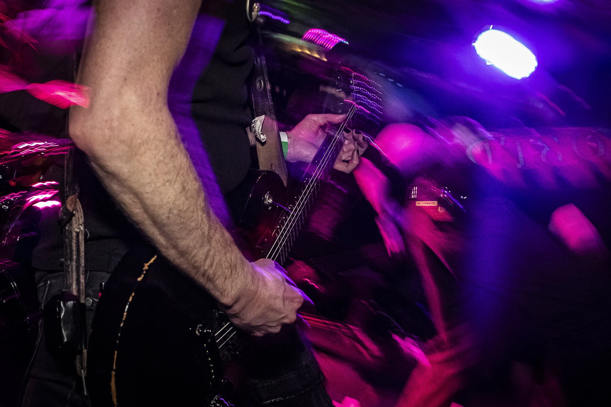 A person playing electric guitar at a music event with purple and pink stage lighting, and blurred crowd in the background.