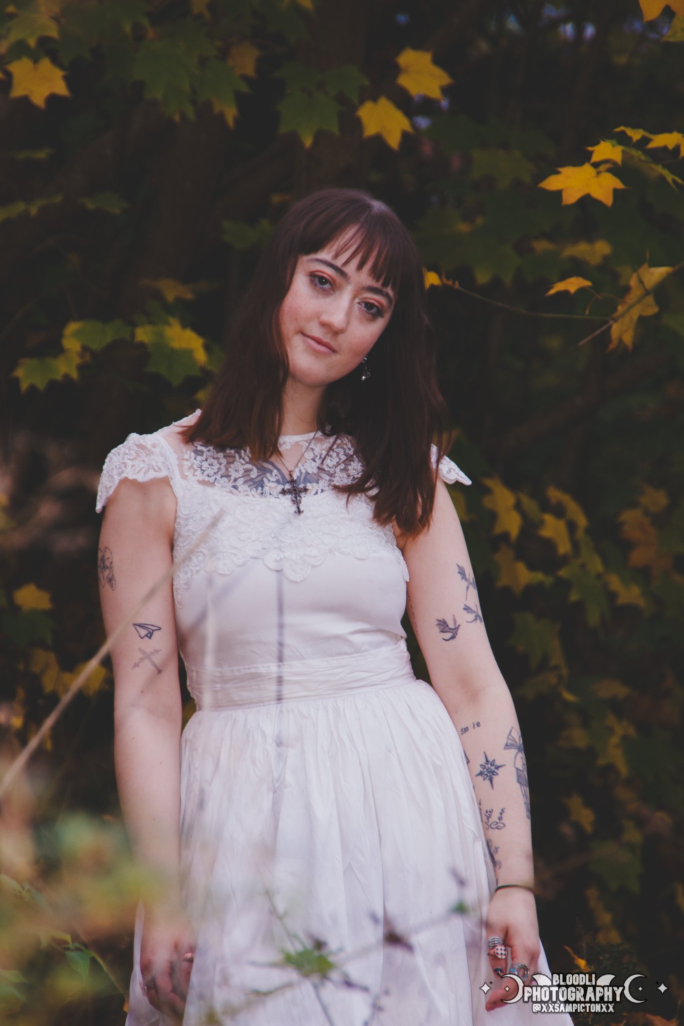 A young woman with brown hair and tattoos on her arms, wearing a white dress, stands outdoors with green and yellow leaves in the background.