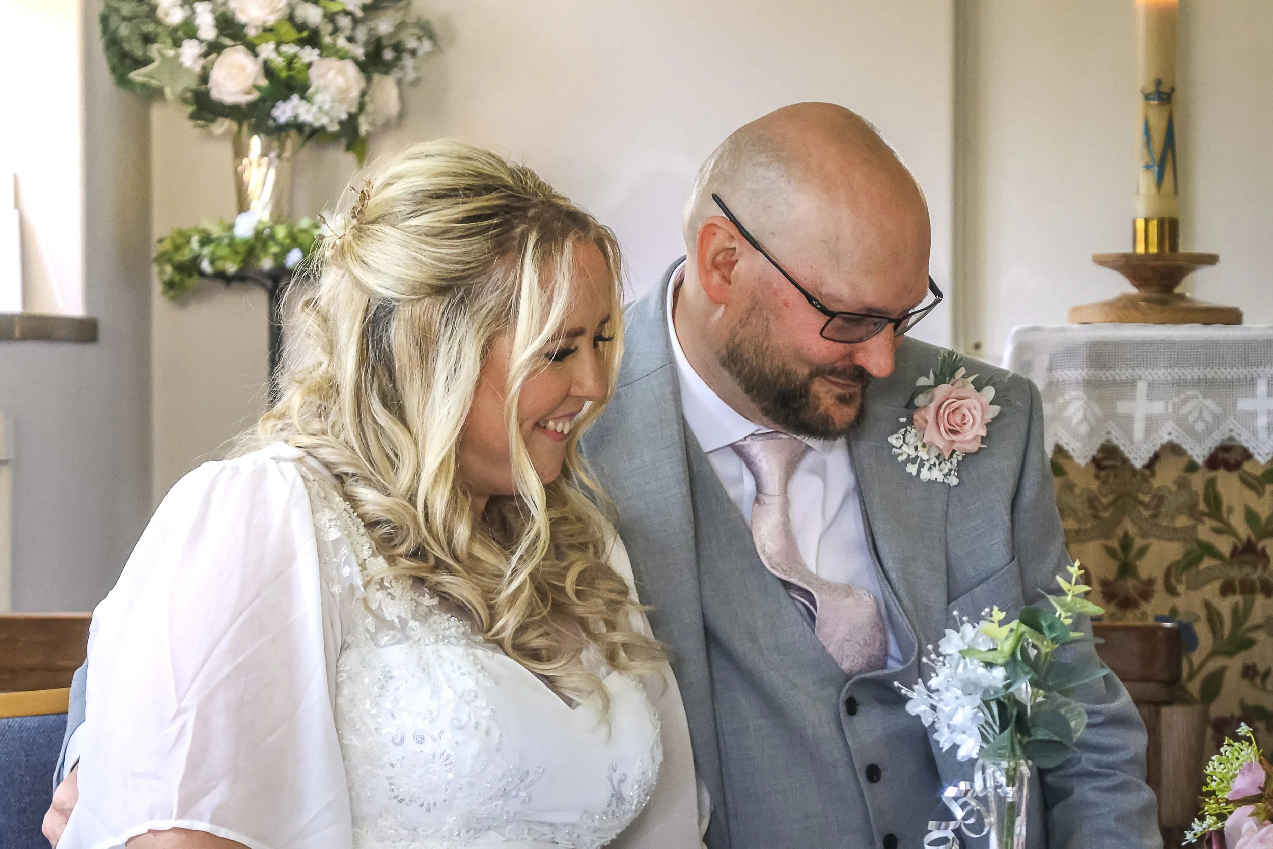 A bride and groom seated in a church during their wedding ceremony, both smiling and looking down, with flowers and religious decorations in the background.