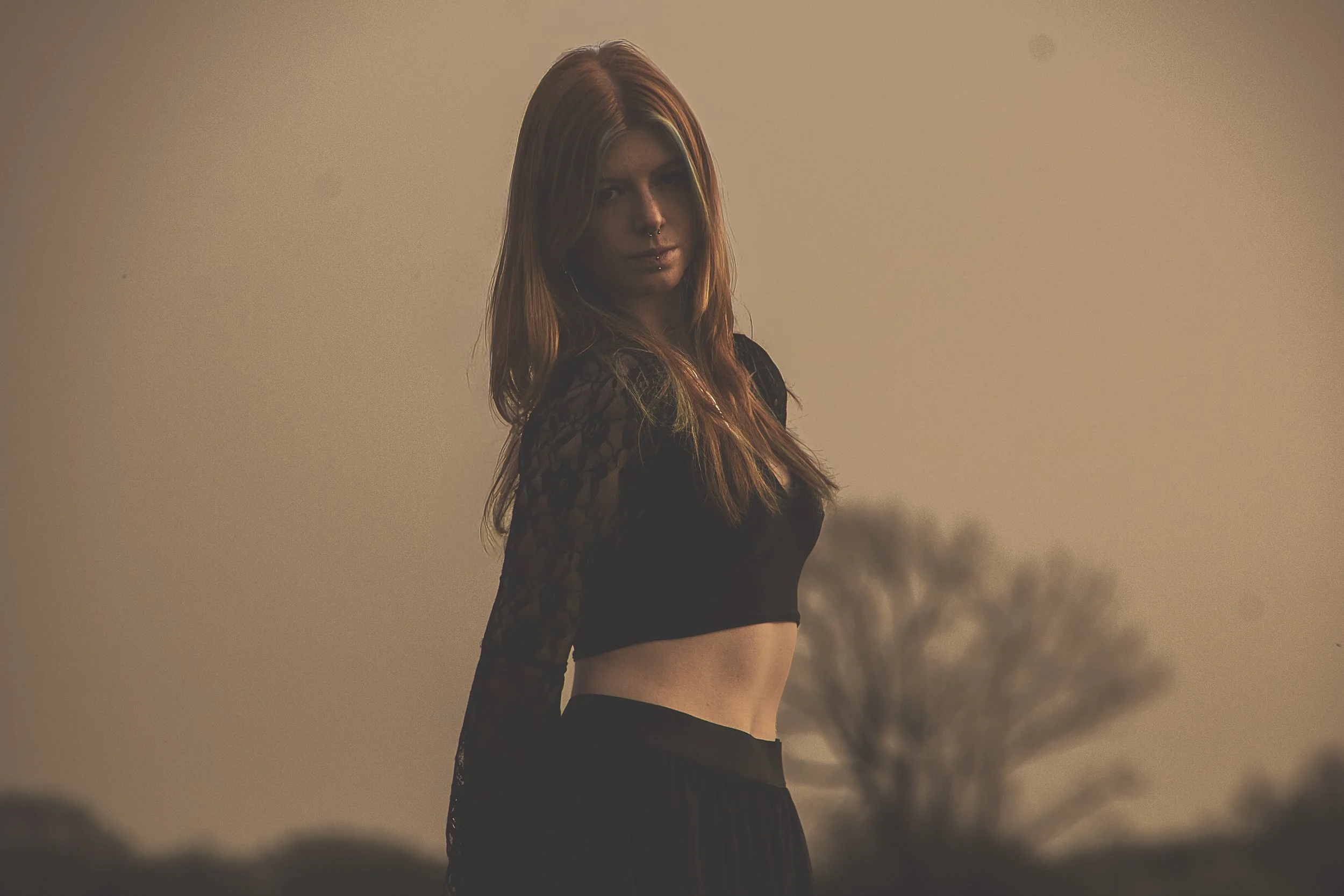A young woman with long red hair standing outdoors during sunset with silhouettes of trees in the background, wearing a black lace crop top and a pleated skirt.