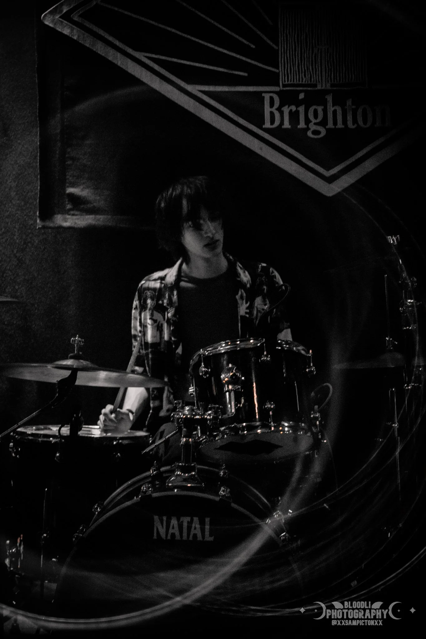 A person playing a drum set on stage with a Brighton poster in the background, in black and white.