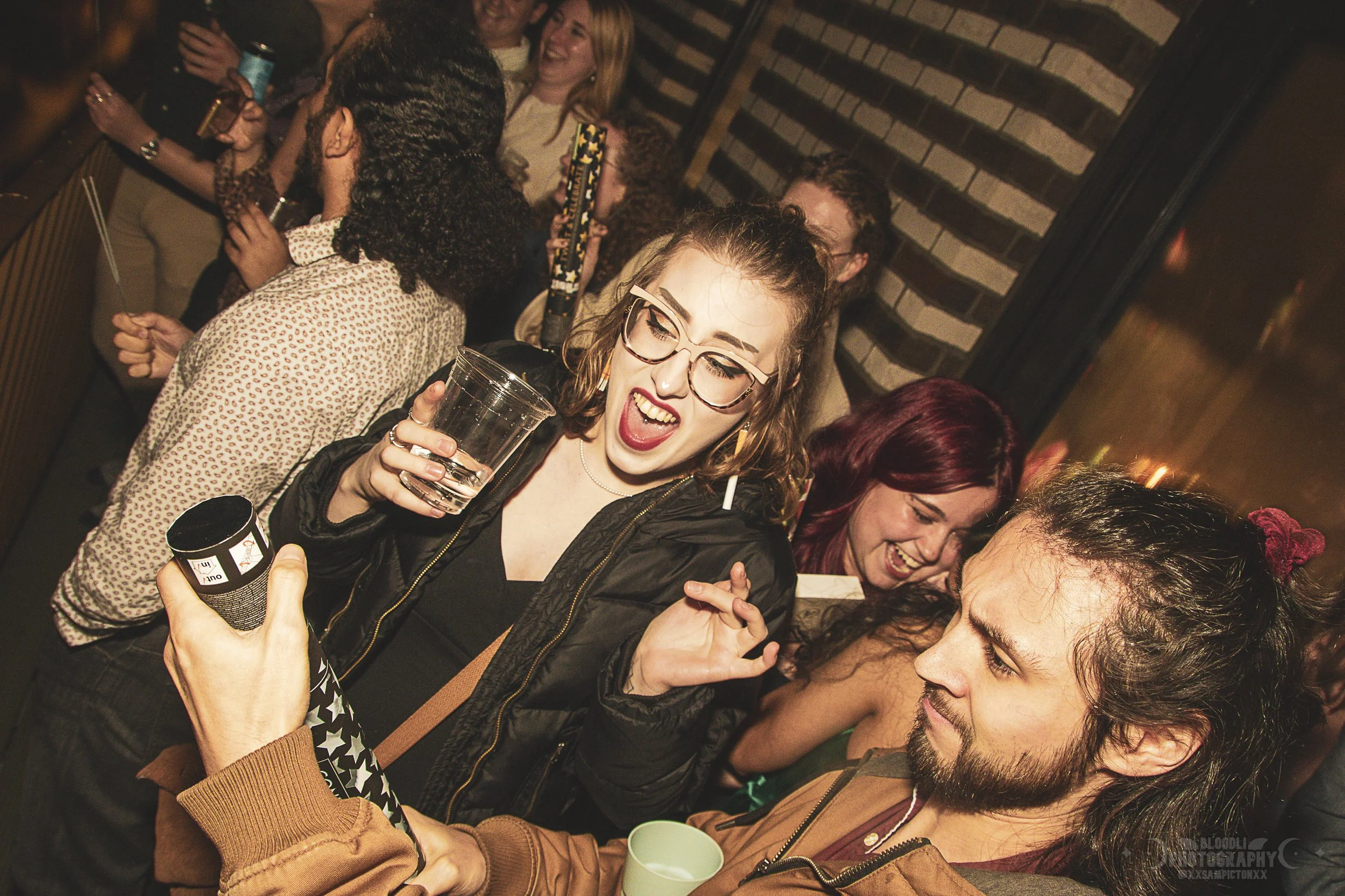 group of people celebrating and socializing at a party or bar, with one woman in the foreground holding a glass of drink and wearing glasses, smiling and talking to a man holding a cup