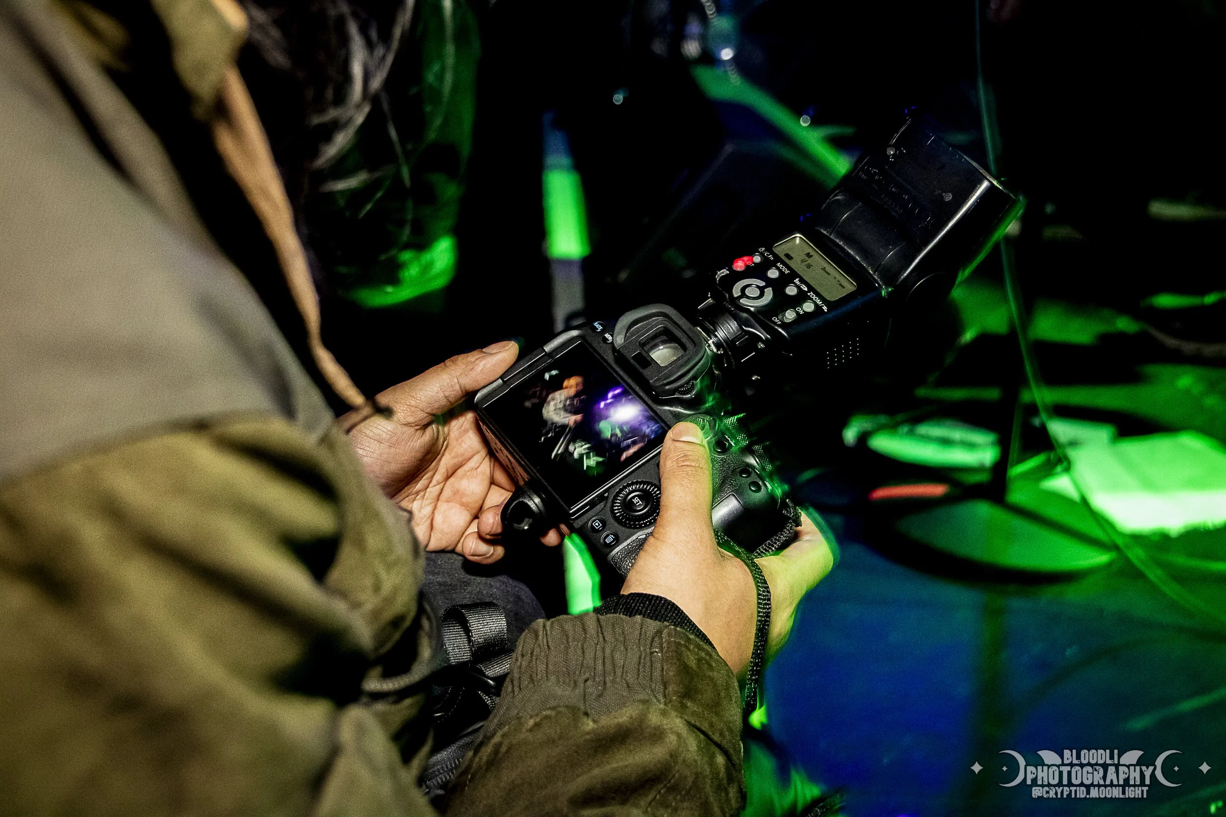 A person in camouflage jacket holding a digital camera, capturing an image of a stage with green and purple lights.