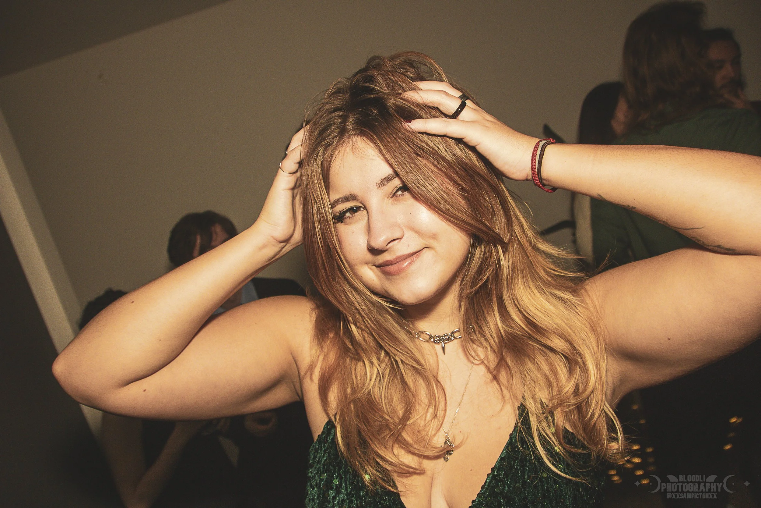 A young woman with long, wavy red hair posing with her hands on her head, smiling and winking at the camera, wearing a dark green dress and jewelry.