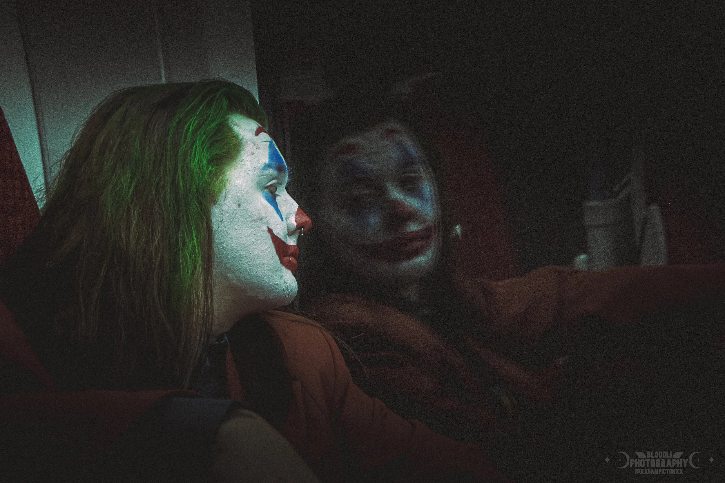 Two women sitting side by side, dressed as clowns, with one wearing white face paint and colorful makeup, the other with dark makeup, in a dimly lit environment.