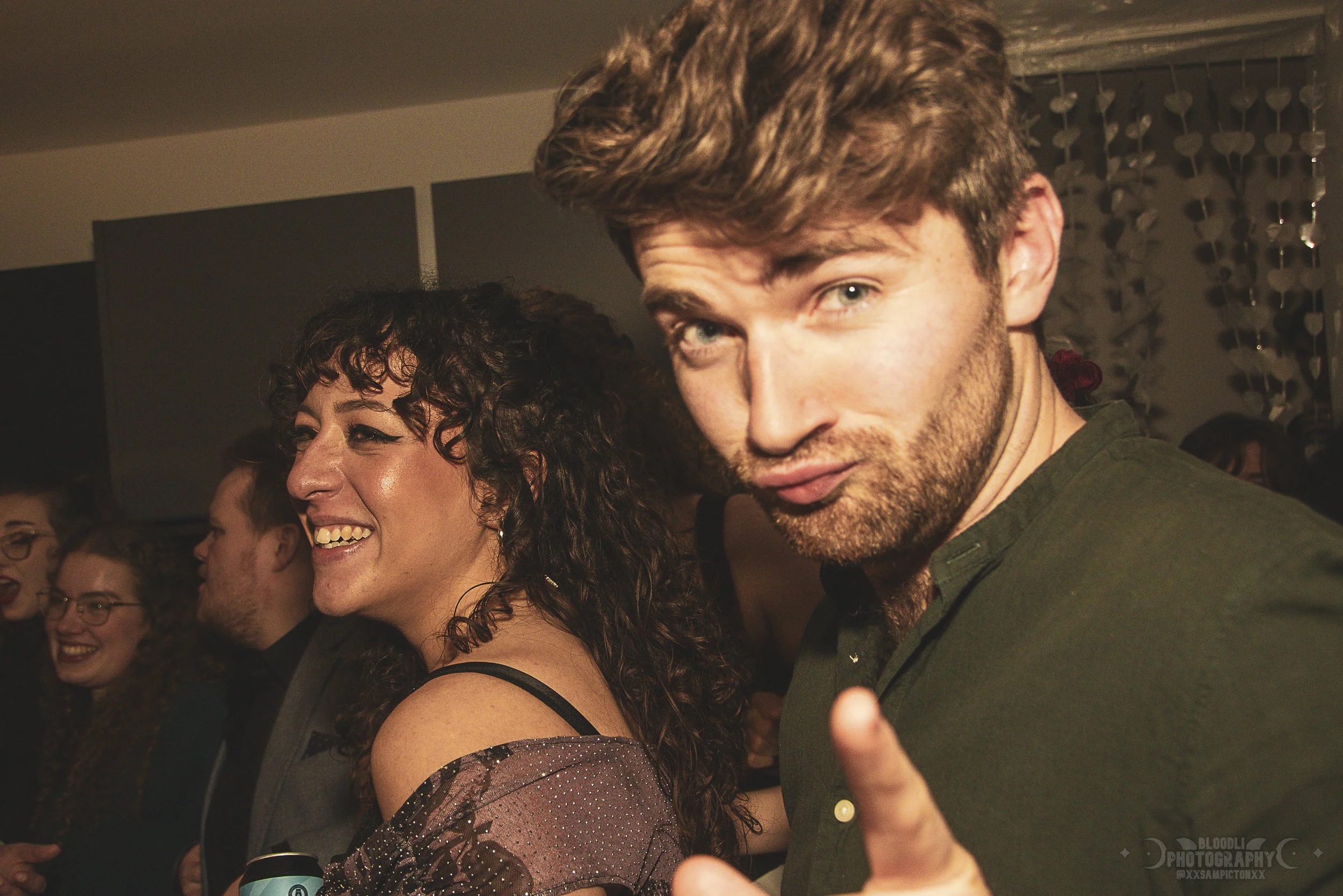 Group of young adults at a party, smiling, with a woman with dark curly hair and a man with light brown hair giving a playful look to the camera.