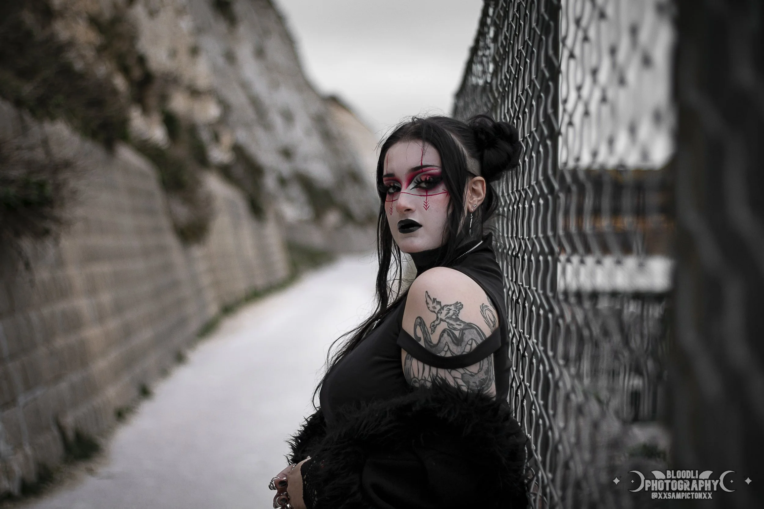 A woman with black hair styled in buns, wearing gothic makeup with red and black face paint, black lipstick, and earrings, leaning against a chain-link fence in an outdoor setting with rocky terrain and cloudy sky.