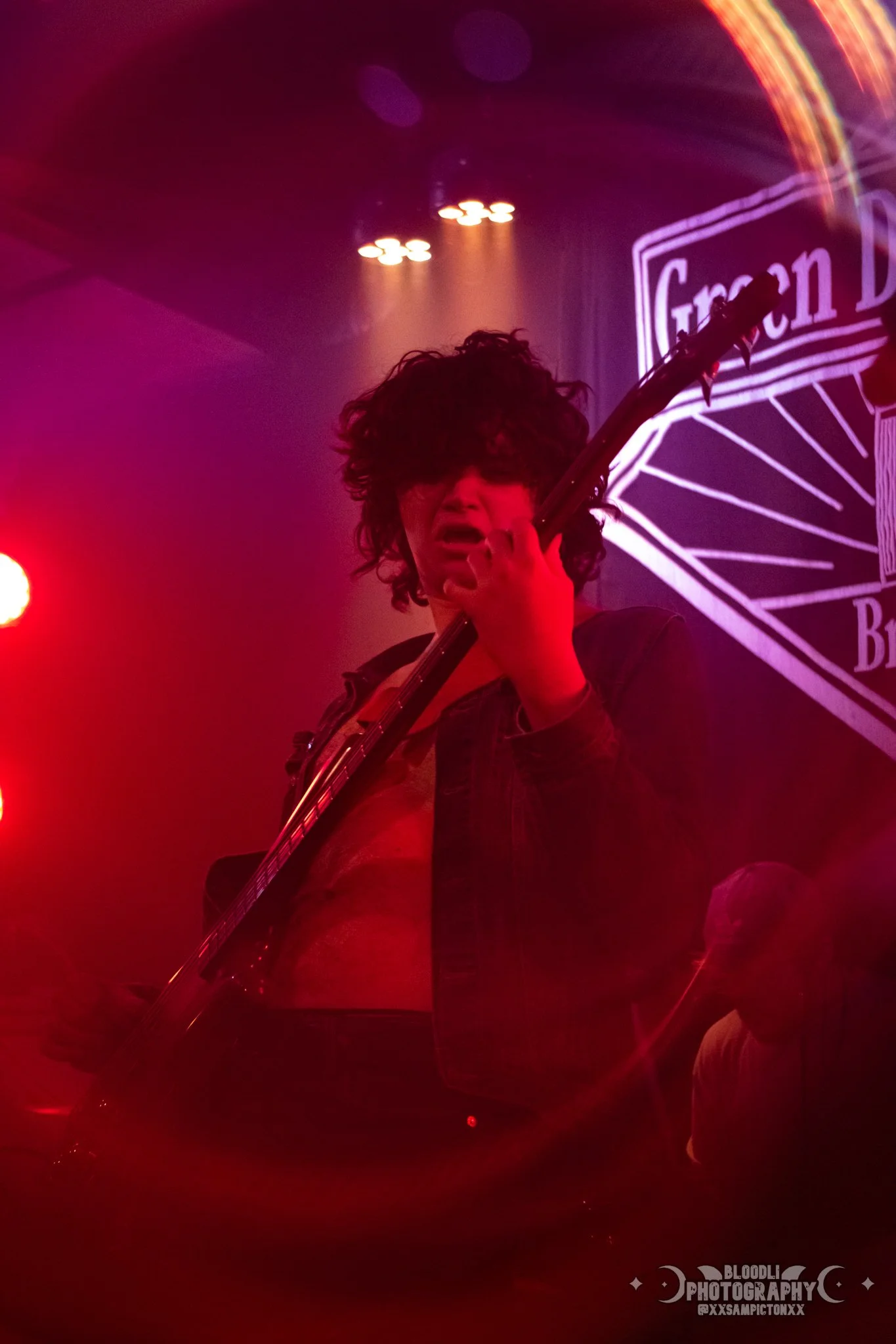 Musician playing guitar at a concert, surrounded by red and purple stage lighting, with a large illuminated sign in the background that reads ''Green D''.