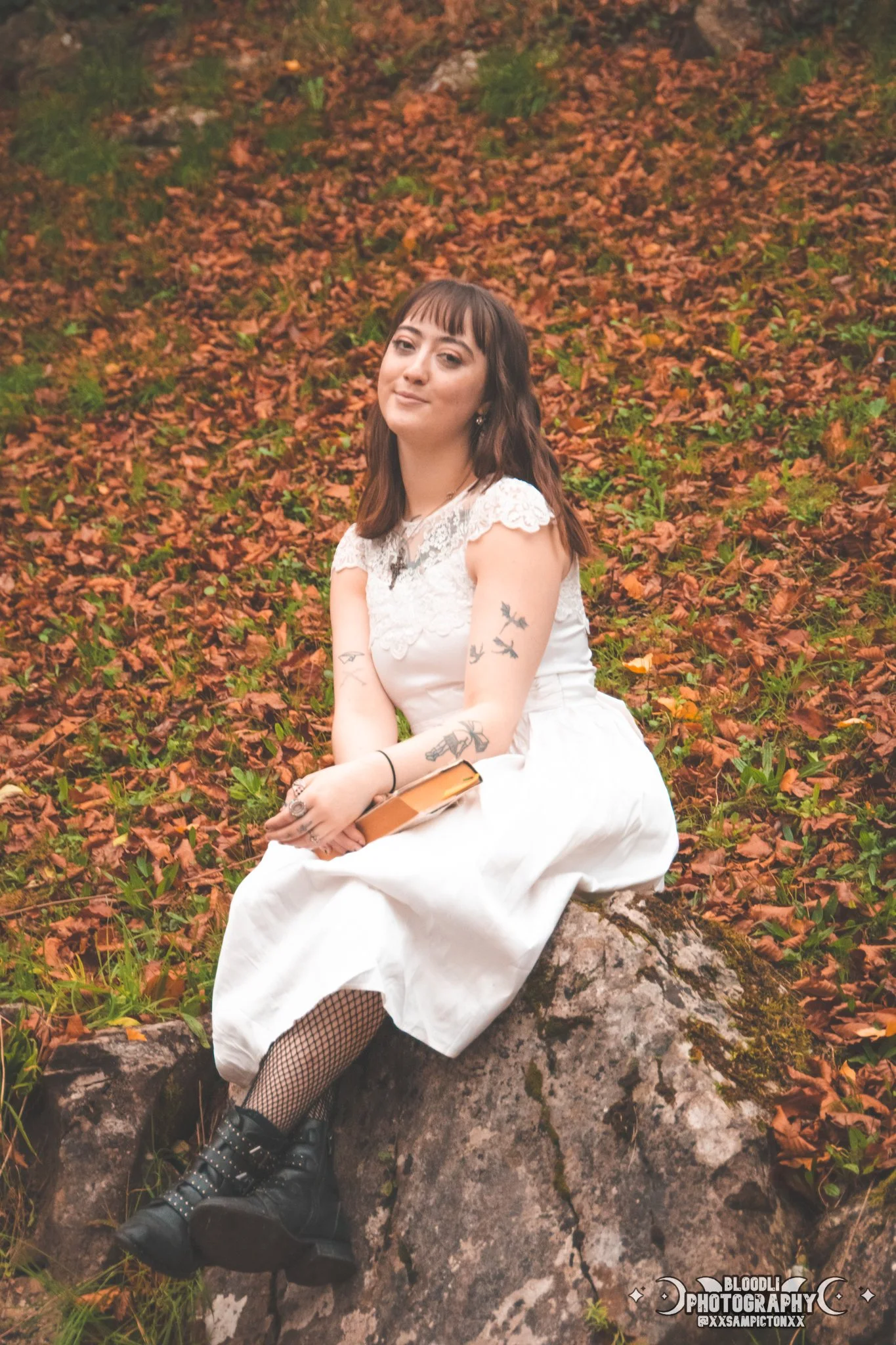 A woman with tattoos sitting on a large rock in a forest surrounded by fallen autumn leaves.