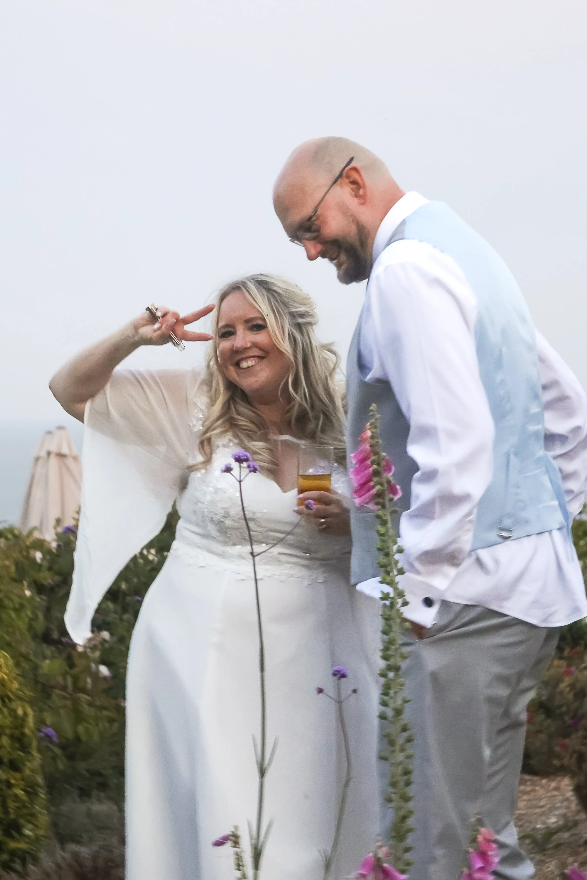 A smiling woman in a white wedding dress and a man in a light-colored vest and shirt with a boutonniere are celebrating outdoors during sunset, surrounded by flowers and greenery.
