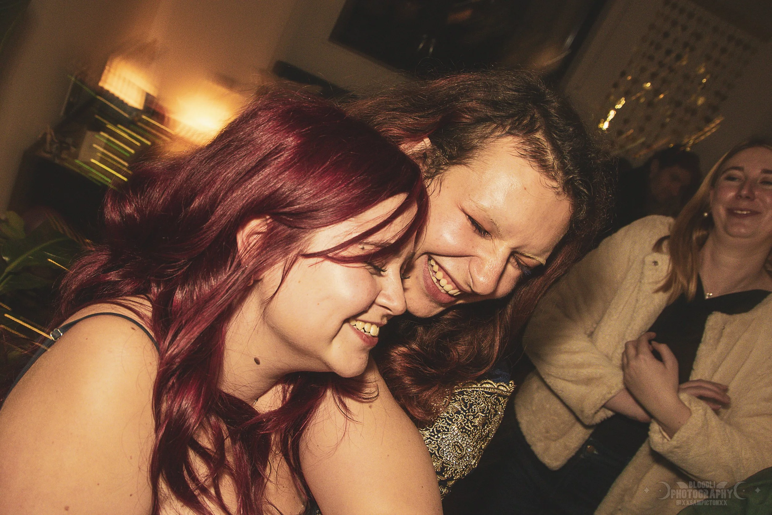 Three women sharing a joyful moment, smiling and leaning their heads together at an indoor gathering.