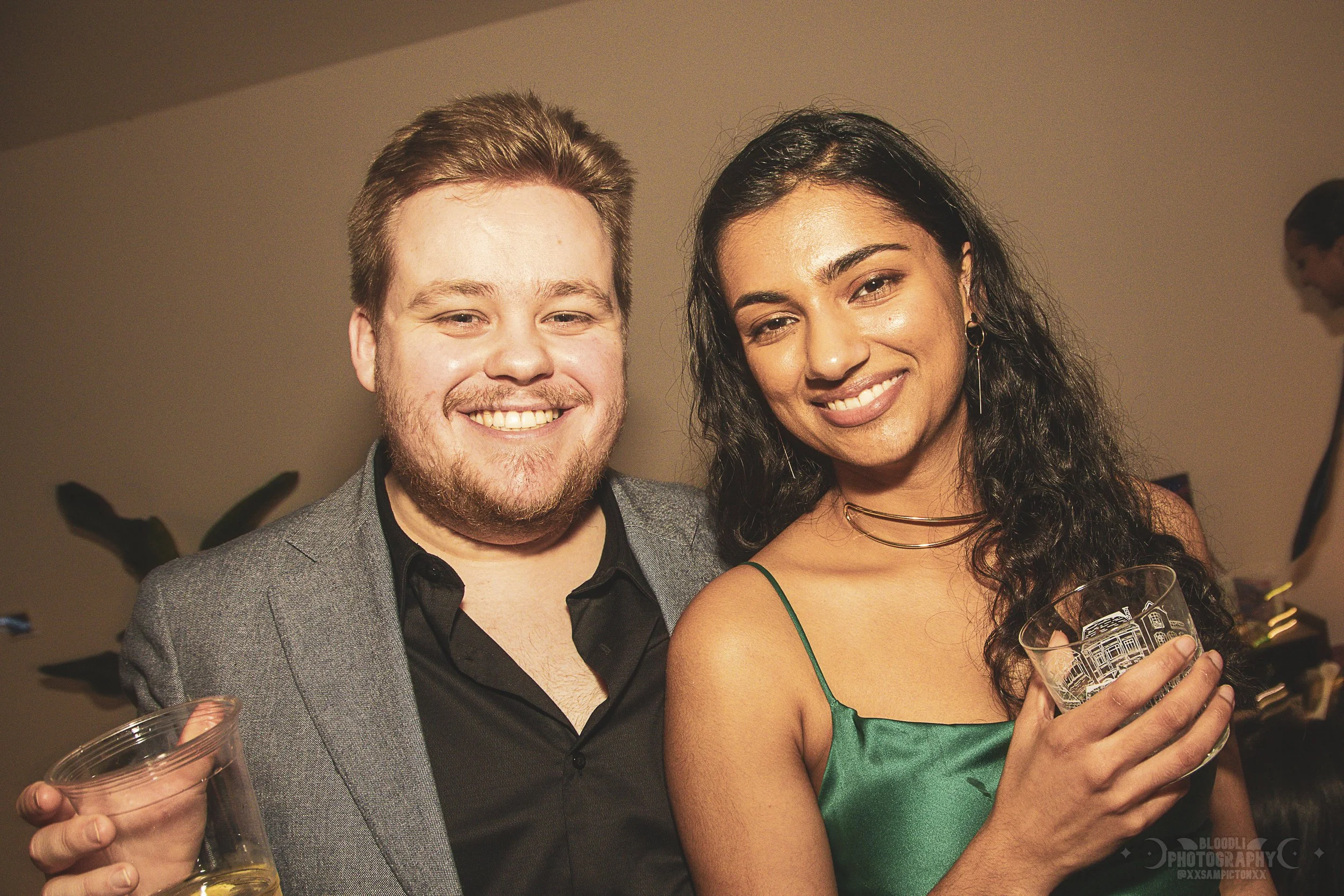 A smiling man and woman posing at a social gathering, holding glasses with drinks.