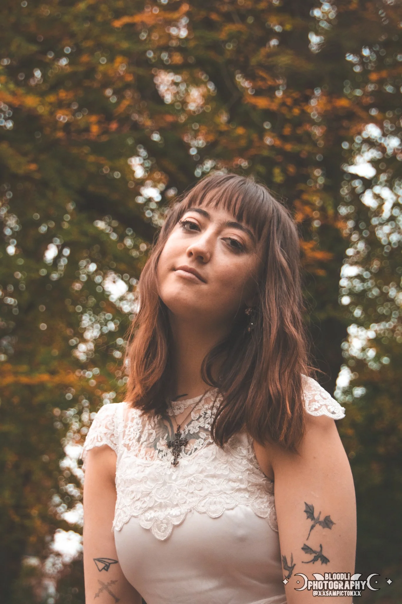 Portrait of a young woman with wavy brown hair and tattoos on her arms, wearing a white lace top, standing outdoors with blurred autumn trees in the background.