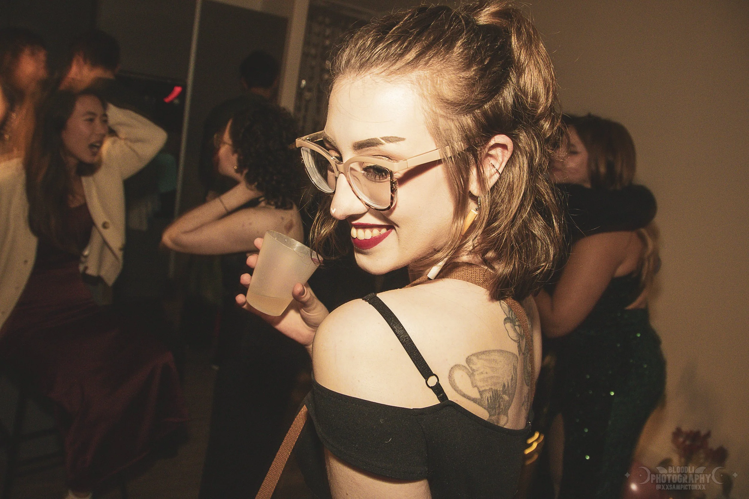 A woman with glasses and short, wavy hair smiling at a party, holding a drink, with other people dancing and socializing in the background.