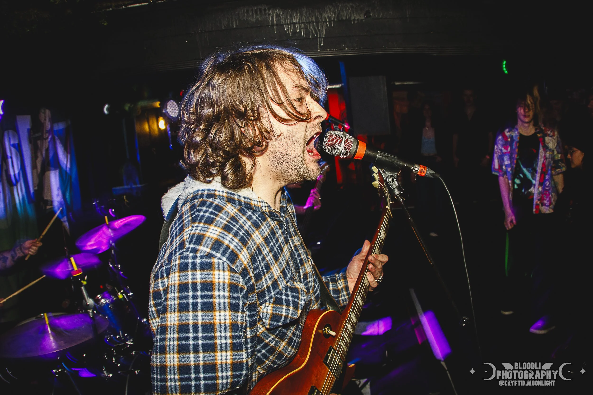 A male musician with shoulder-length hair and a plaid shirt singing into a microphone while playing an electric guitar at a live music event.