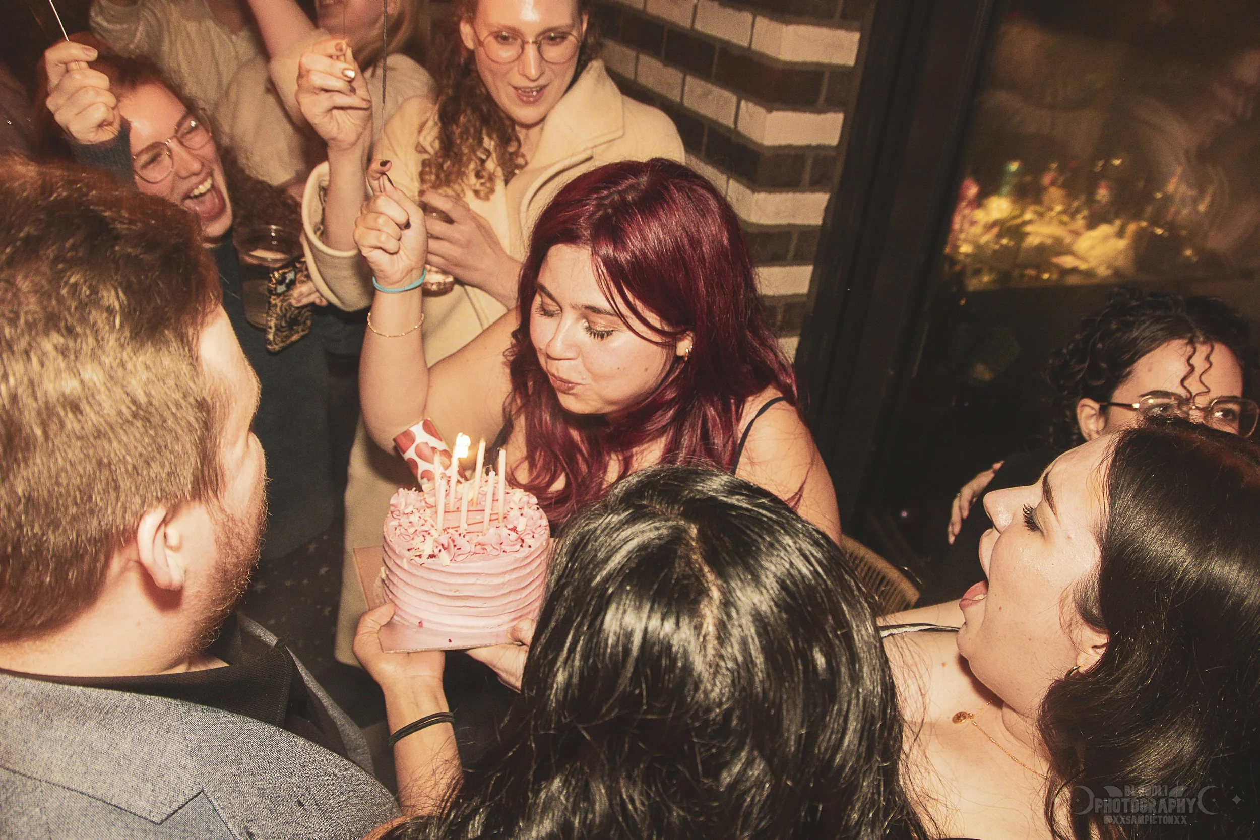 A woman with red hair is blowing out candles on a pink birthday cake while surrounded by a group of friends celebrating.