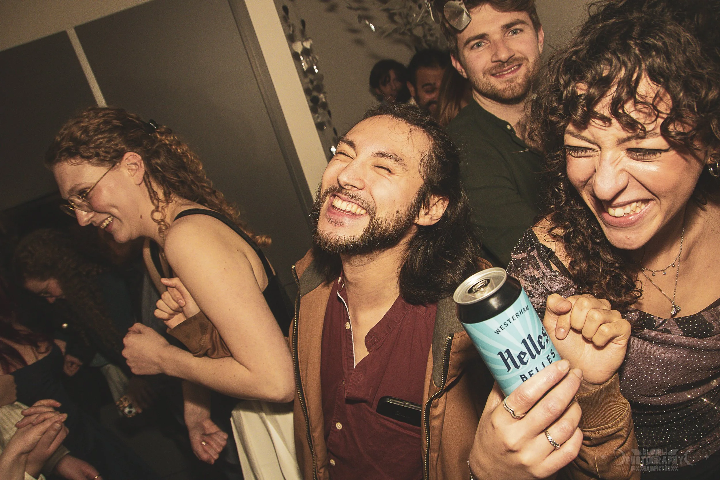People at a crowded party laughing and enjoying themselves. One man in the foreground is holding a can of beer and smiling with his eyes closed. Women around him are also smiling and laughing, creating a lively atmosphere.