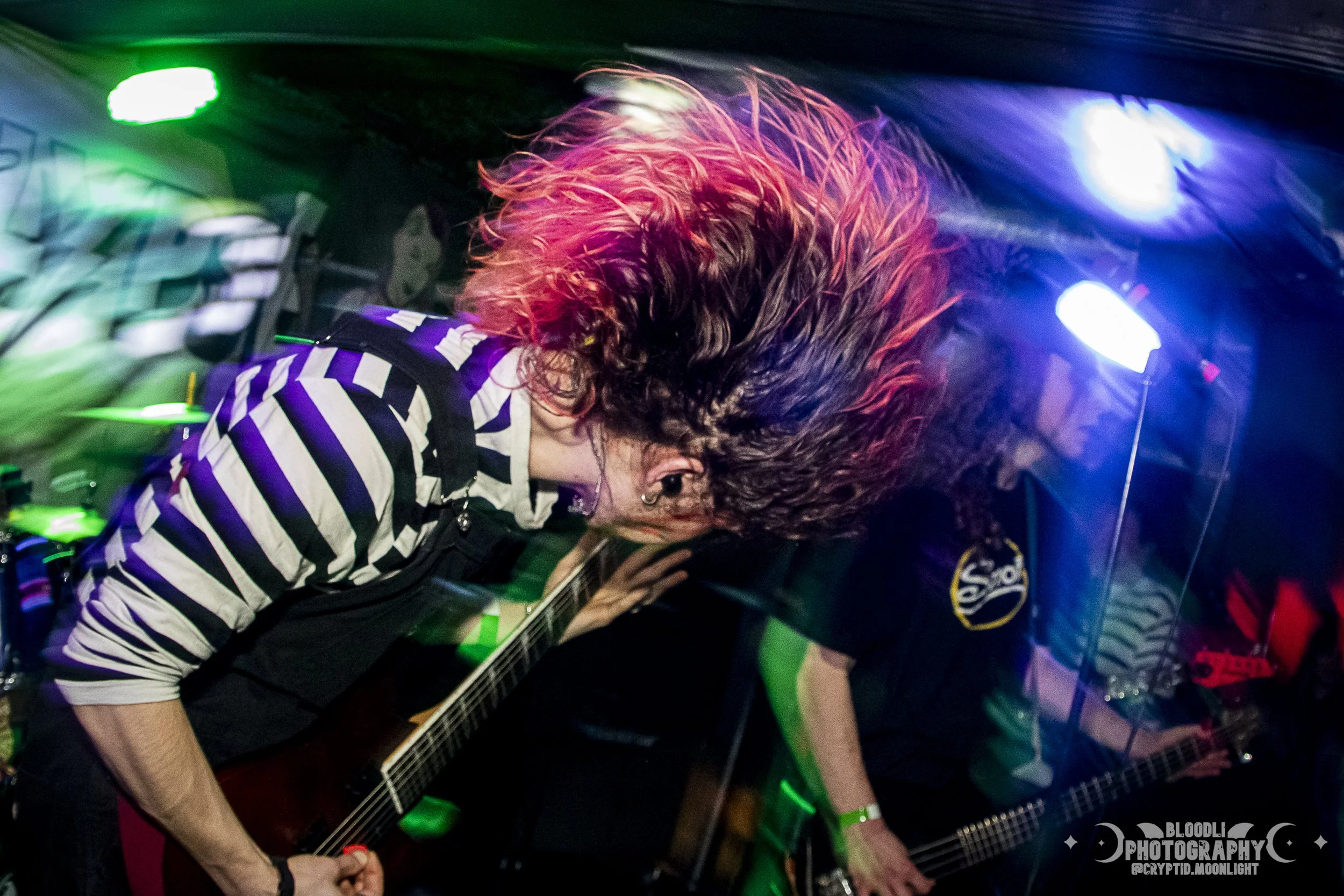 Musician with long flowing hair playing guitar on stage with colorful lights in the background.