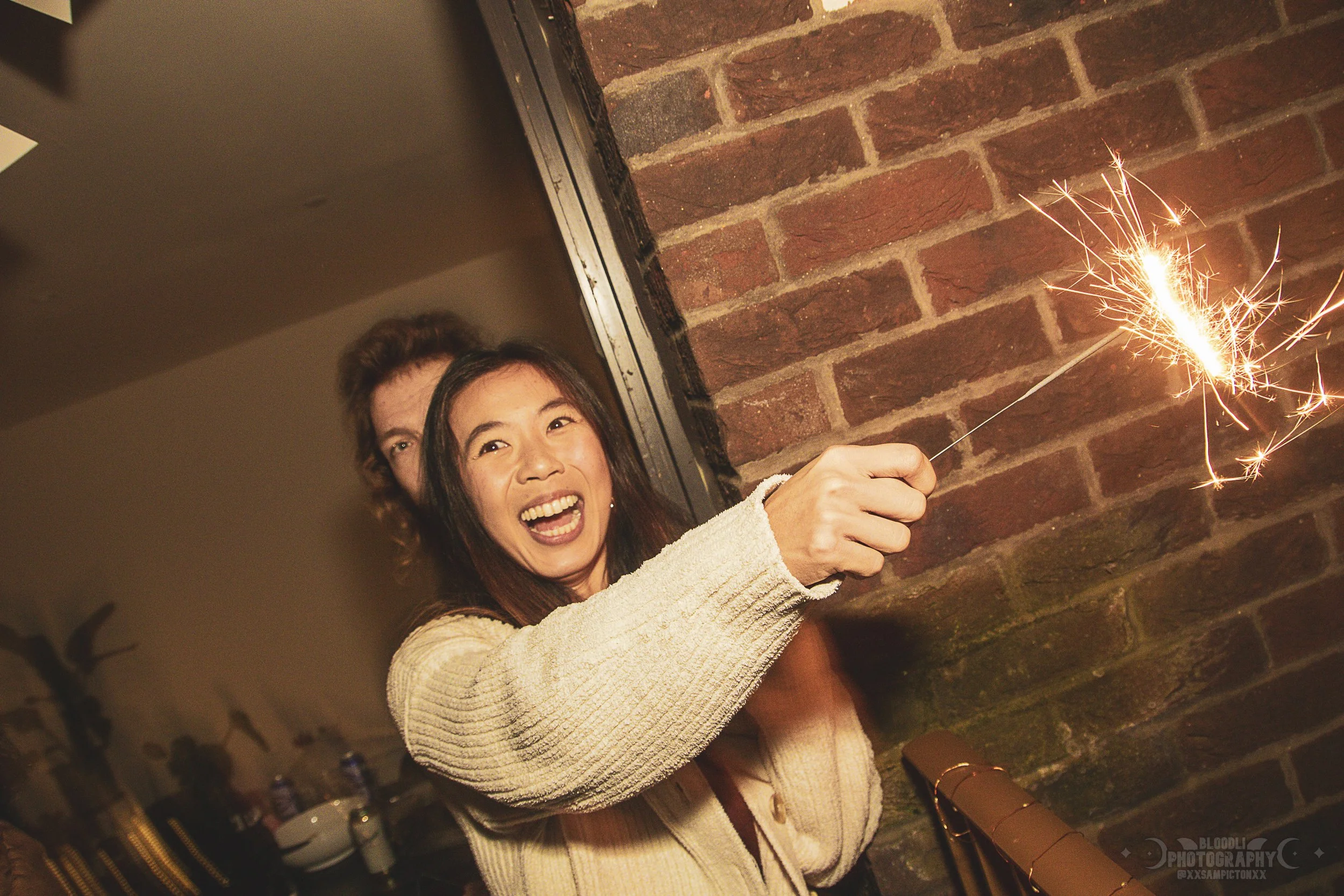 A woman smiling and joyfully holding a lit sparkler while celebrating, with a brick wall in the background.