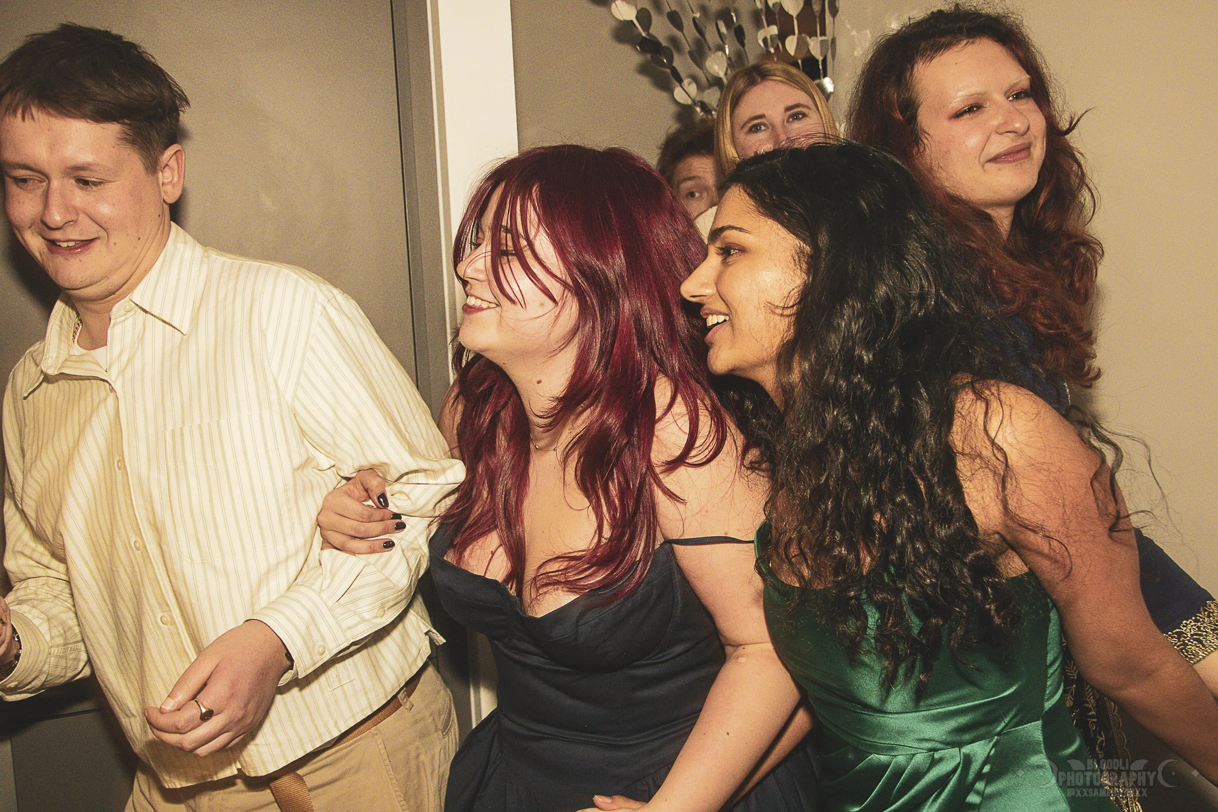 Group of people at a party or social gathering, standing close together, smiling, dressed in semi-formal attire.