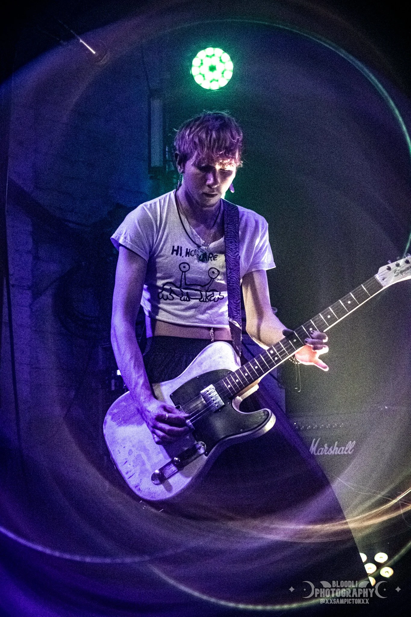 A young person playing an electric guitar on stage with purple and green lighting, wearing a white T-shirt with a cartoon crab drawing and black pants, with a Marshall amplifier in the background.