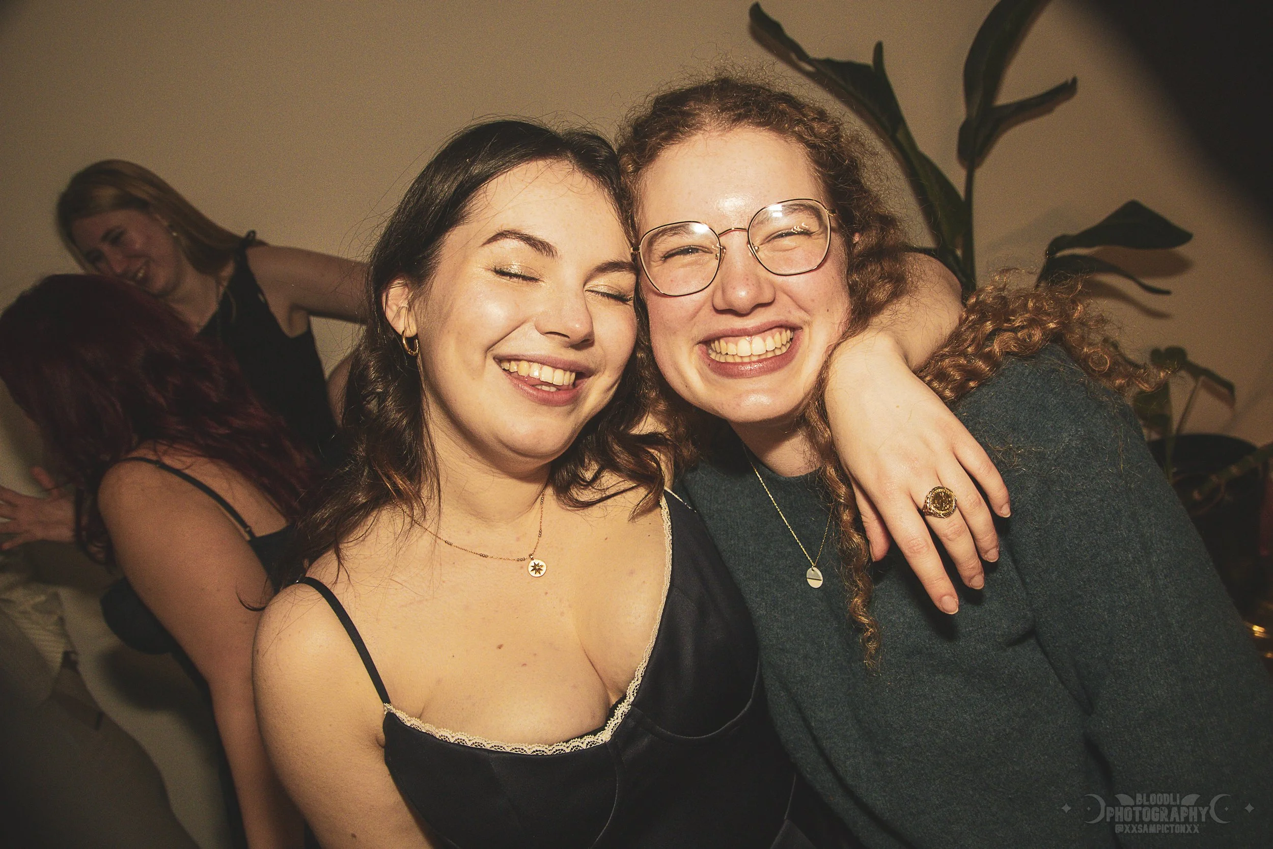 Two women smiling and hugging at a party, with two other women dancing in the background.