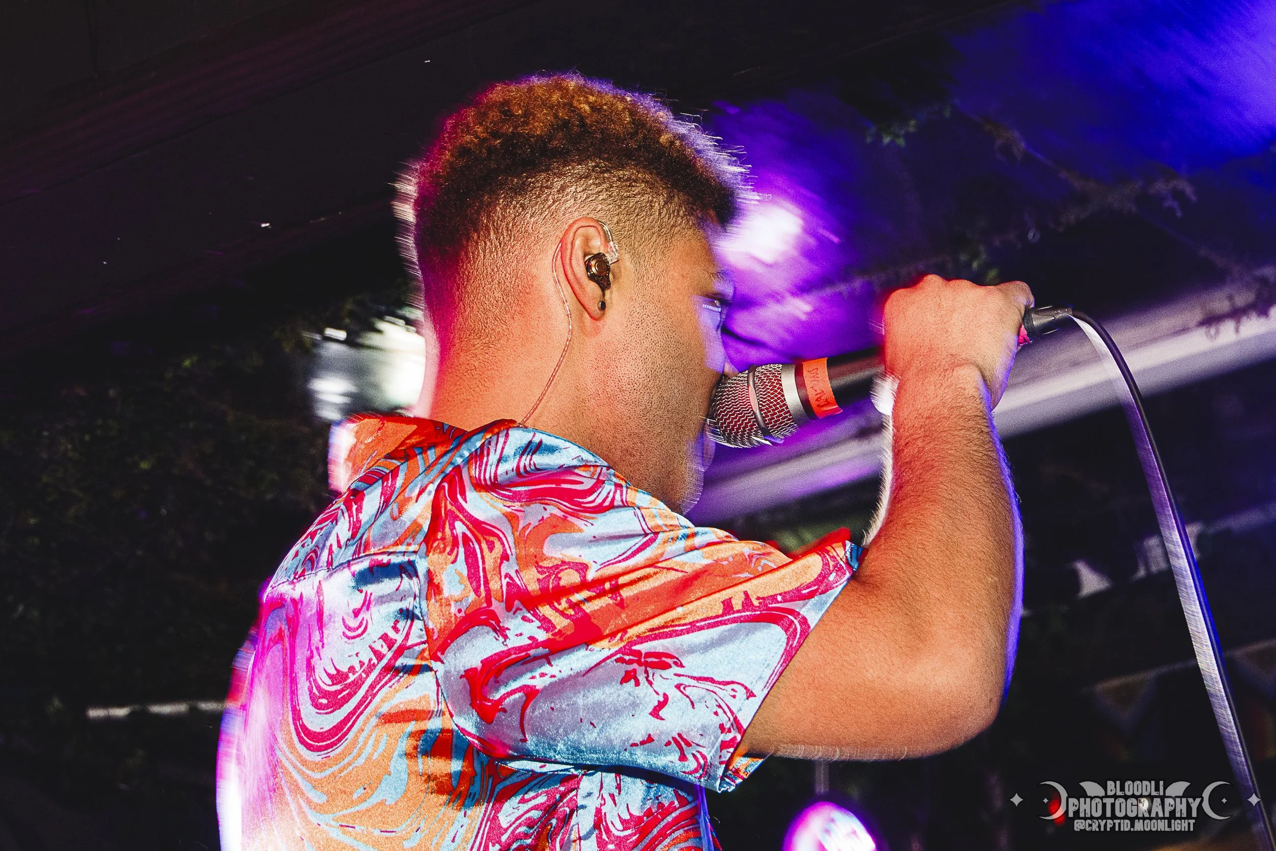 A young man singing into a microphone on stage, wearing a colorful, patterned shirt and in-ear monitors, with stage lighting in the background.