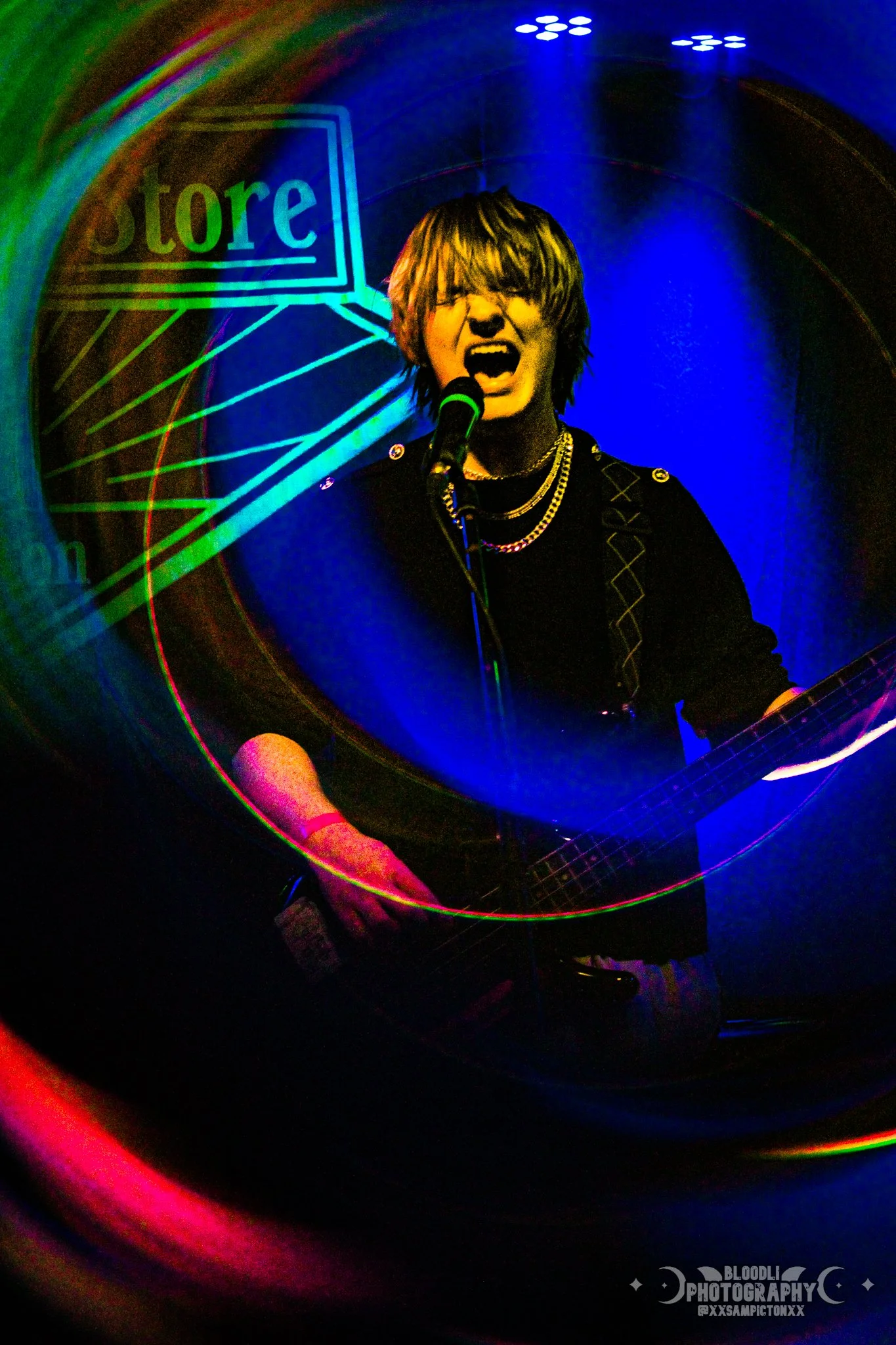 A person singing into a microphone and playing a guitar on a stage with blue lighting and neon signs, creating a colorful, circular light effect.