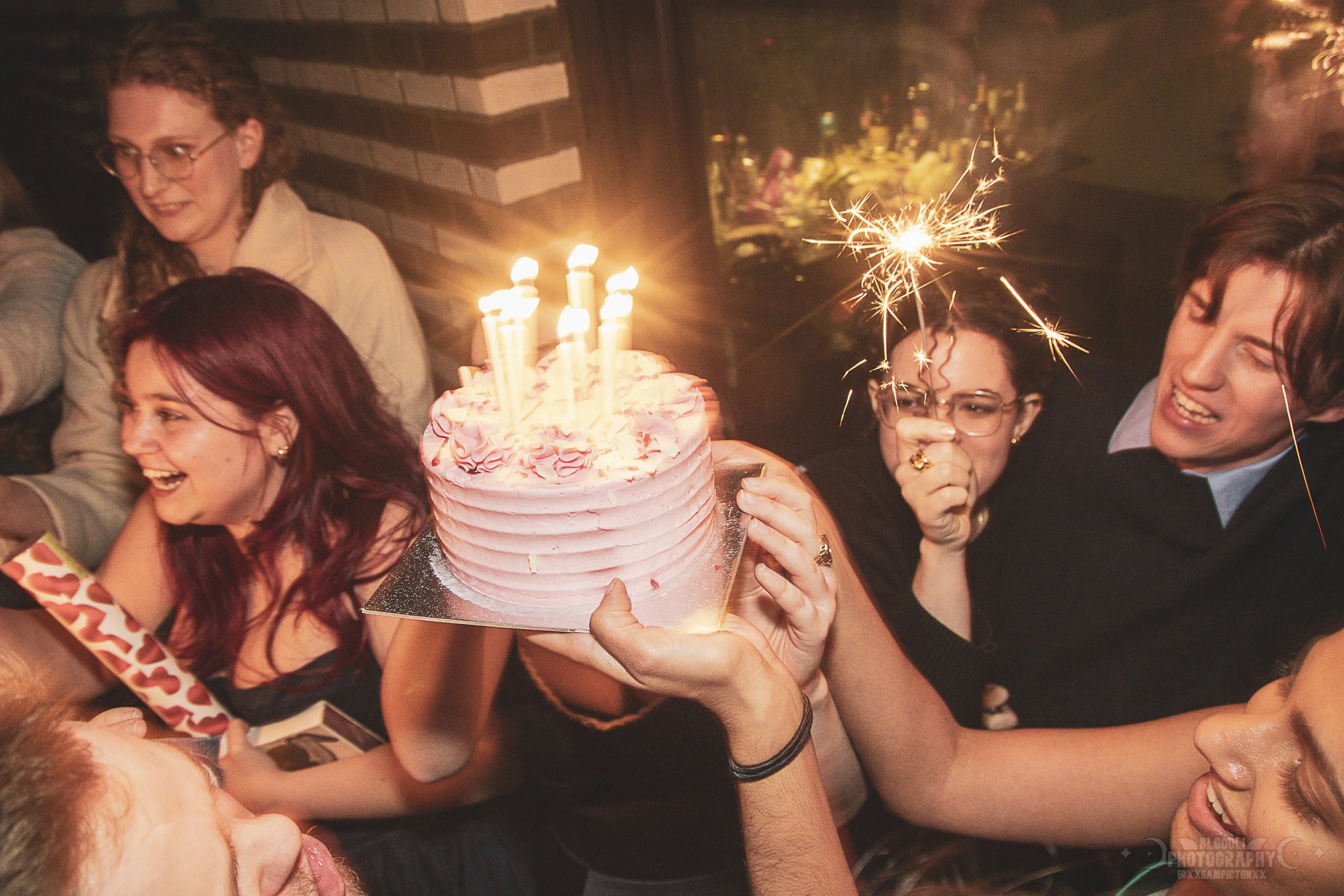 Group of people celebrating birthday with cake, candles, and sparklers in a festive setting.