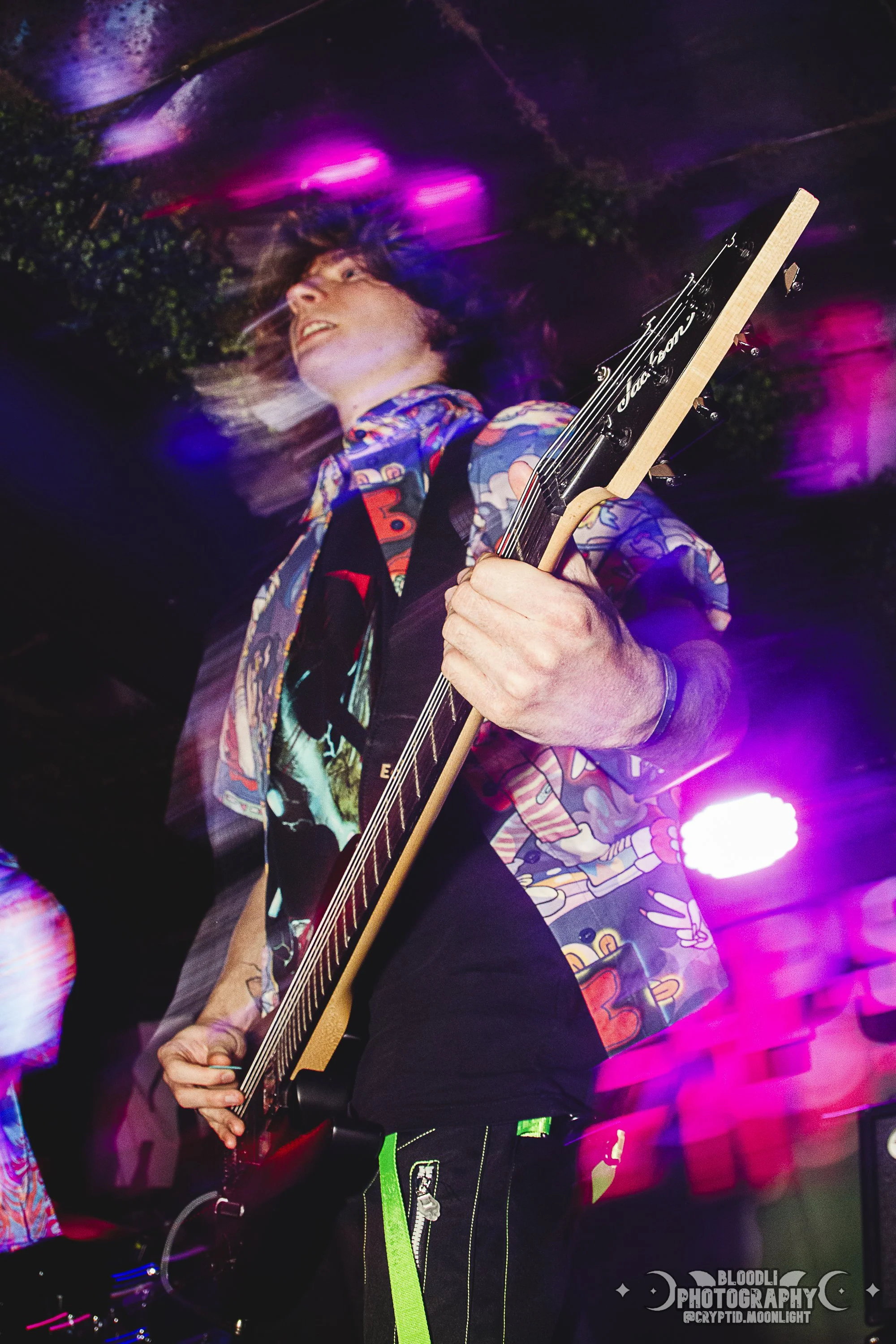 A person playing an electric guitar at a concert, wearing a colorful, patterned shirt and black pants with neon green accents, with pink and purple stage lights in the background.