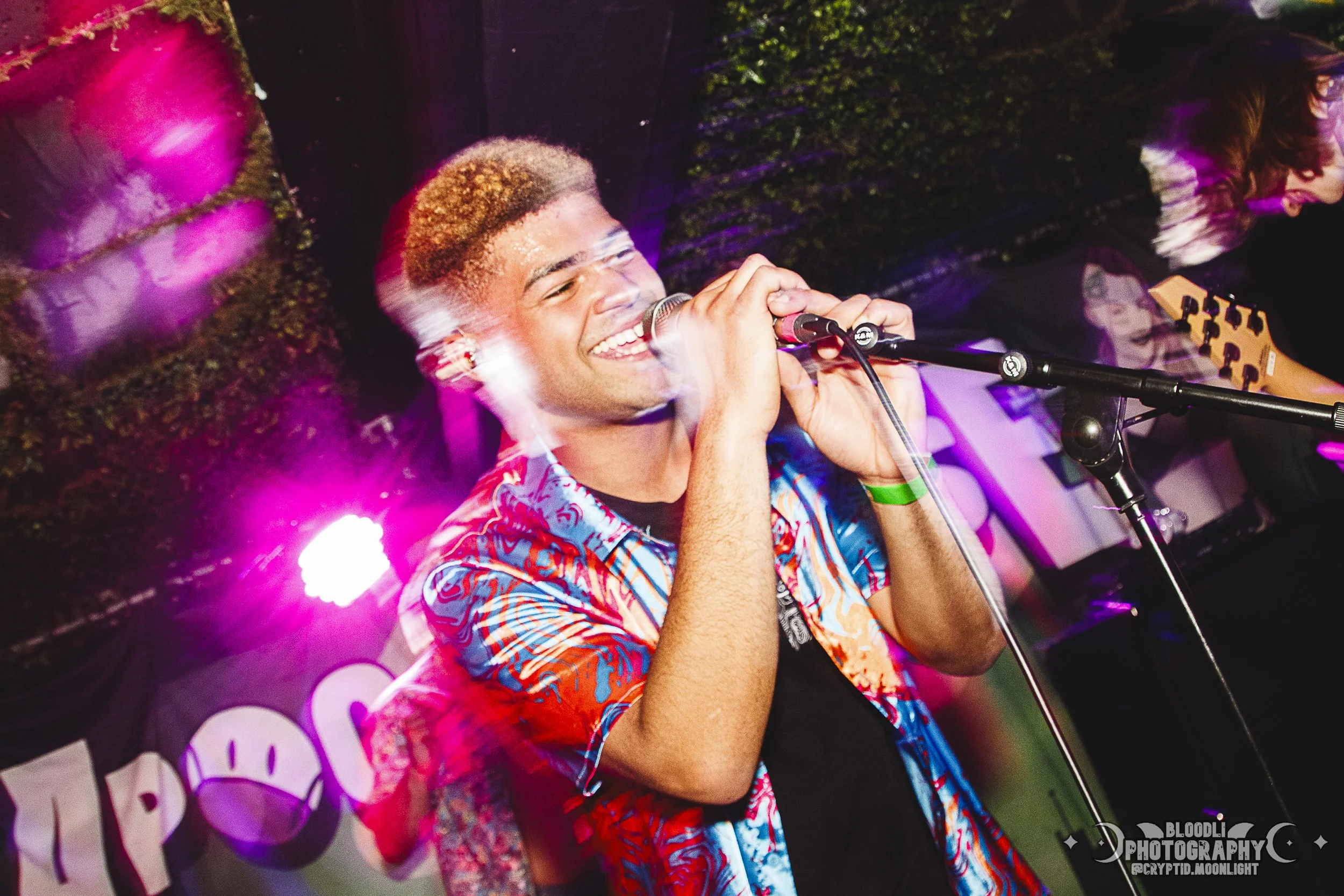 A young man with short curly hair singing into a microphone on stage, wearing a colorful patterned shirt with pink, orange, and blue tones, smiling with closed eyes, in a lively indoor concert setting with pink and purple stage lights.