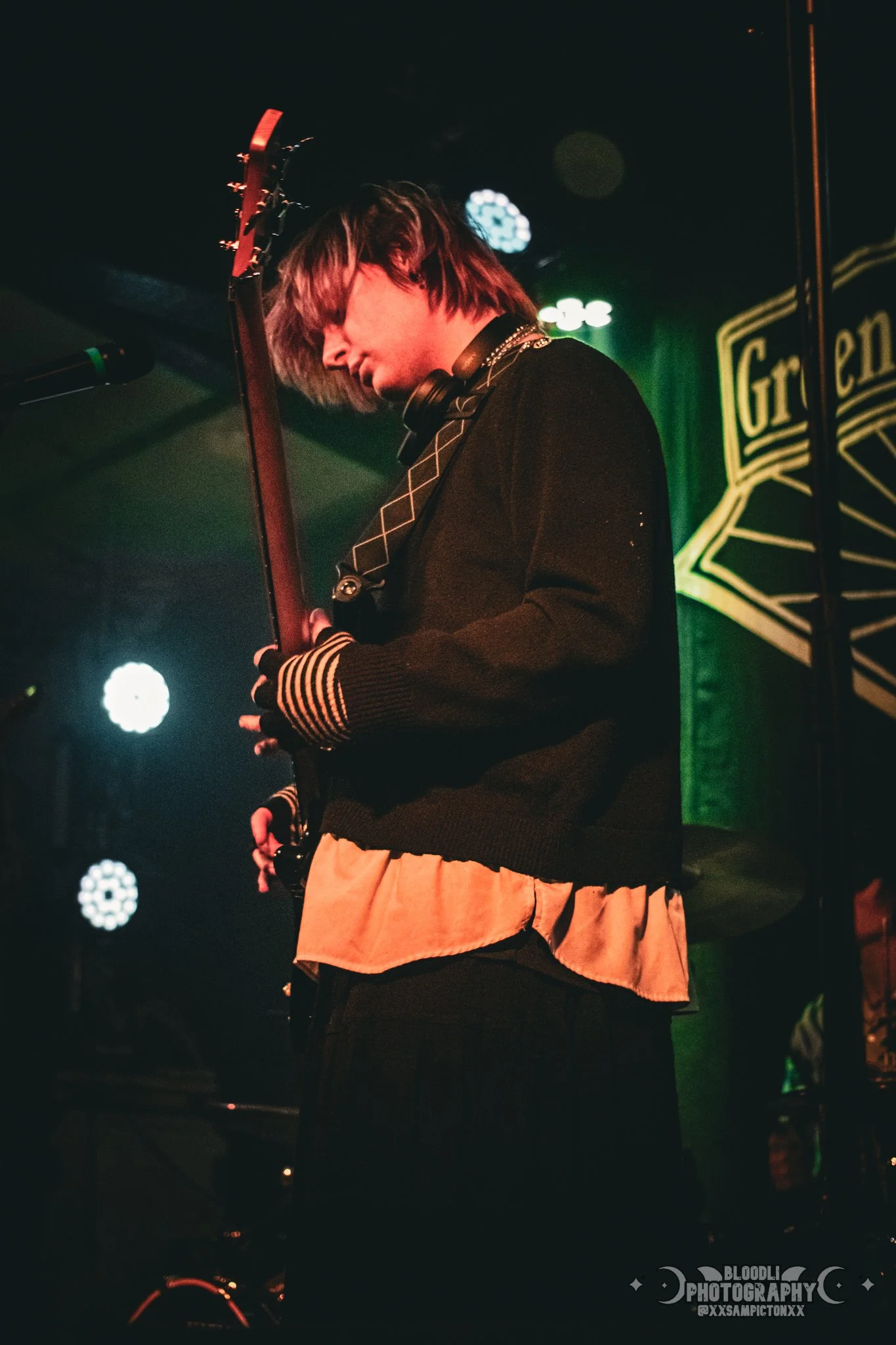 A young musician playing an electric guitar on stage in a dimly lit venue with colorful stage lights and a green backdrop.