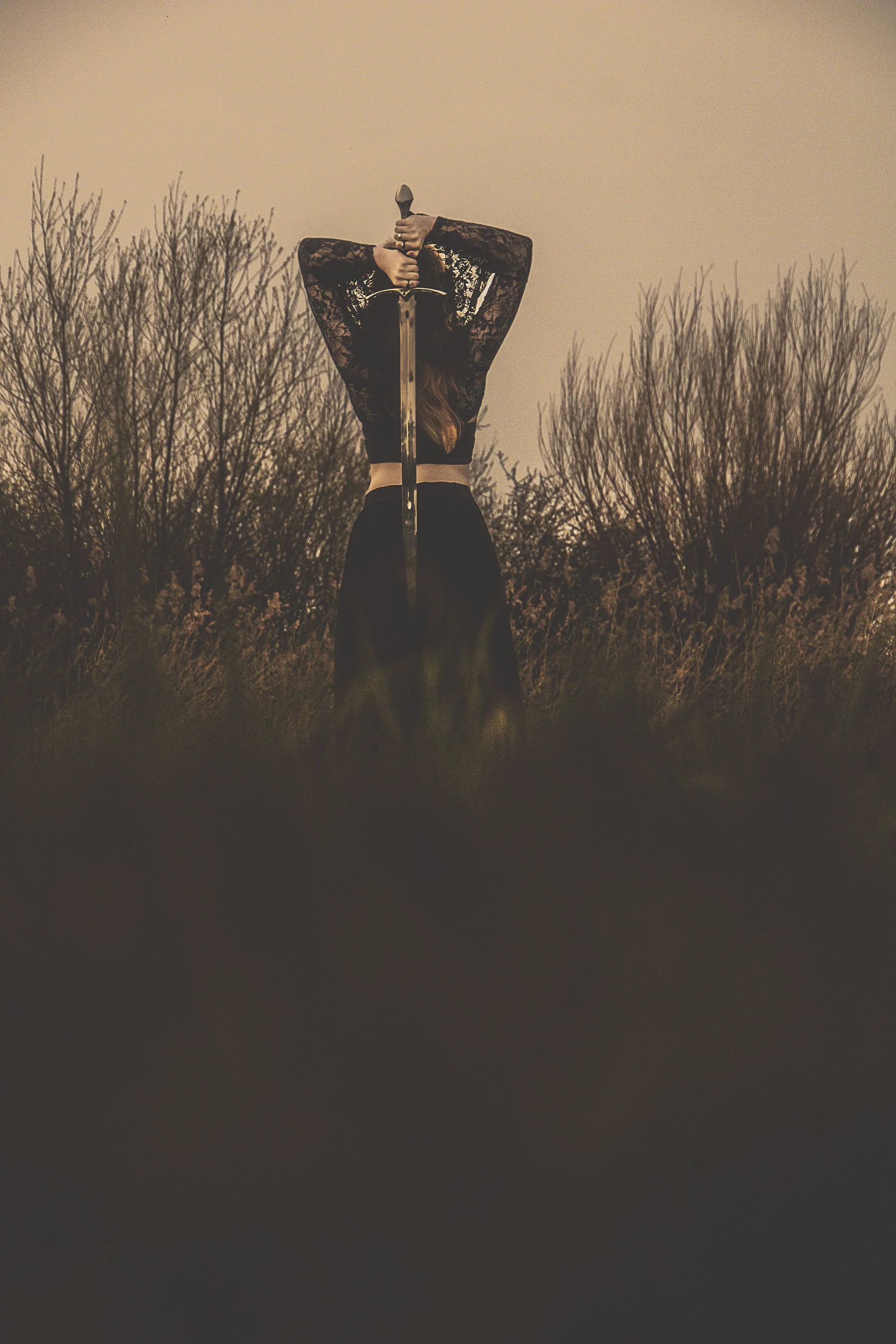 A woman standing outdoors on a grassy field at dusk, holding a sword upright in front of her with both hands overhead, with leafless trees in the background.