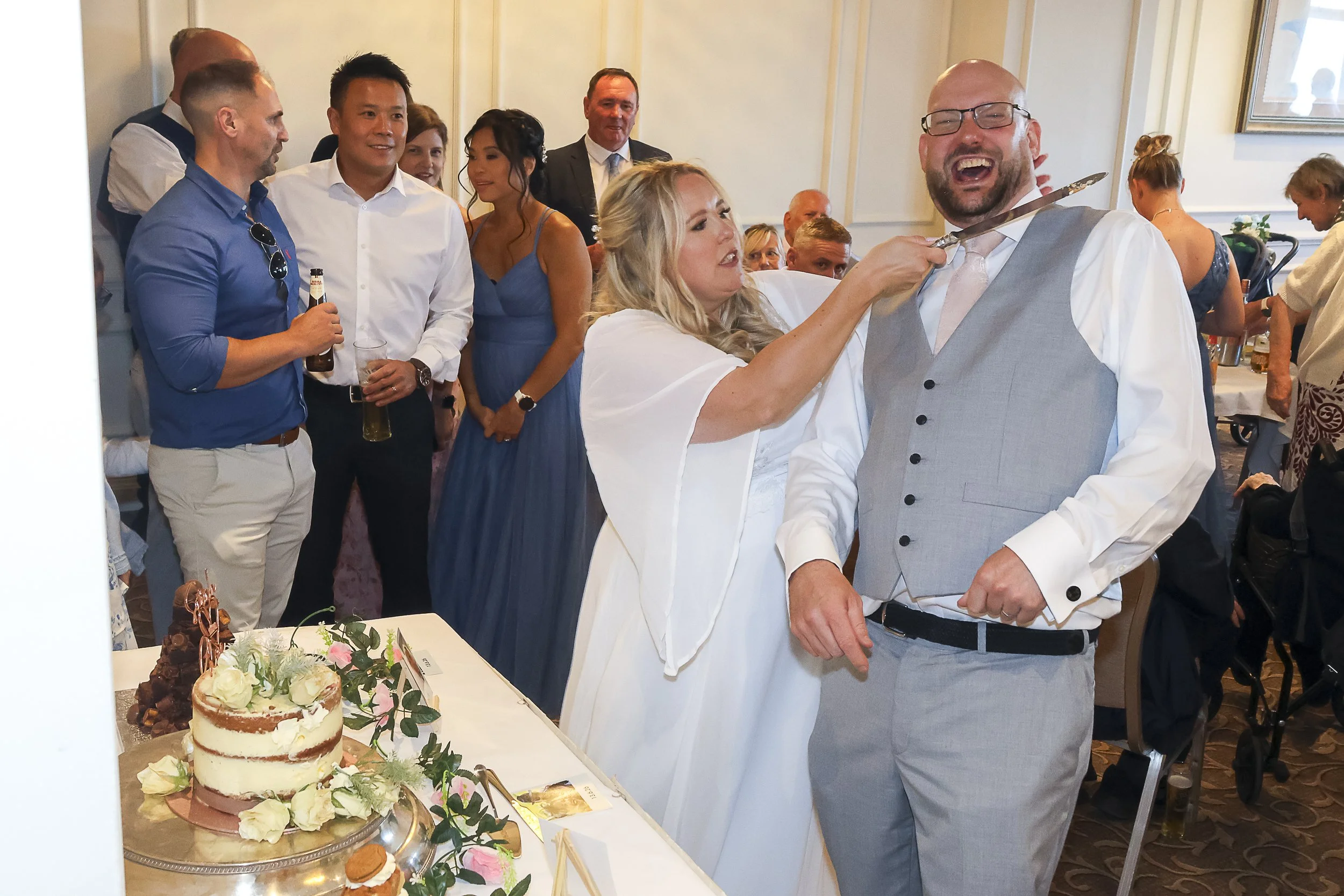 A joyous wedding reception scene with a bride and groom in the foreground, surrounded by guests, including a man with glasses holding a cake knife, ready to cut a wedding cake decorated with flowers.