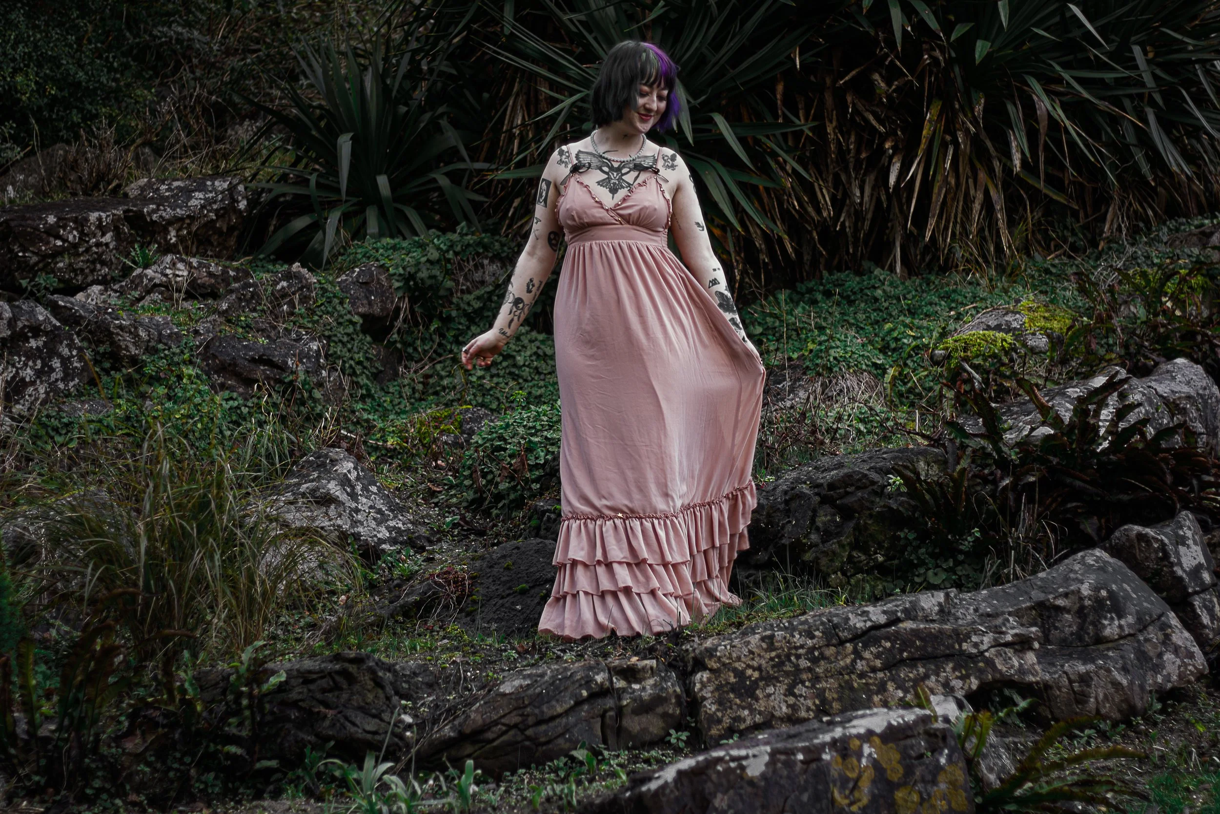 A woman with dark hair and tattoos on her arms is wearing a long, pink dress and standing outdoors among rocks and greenery.