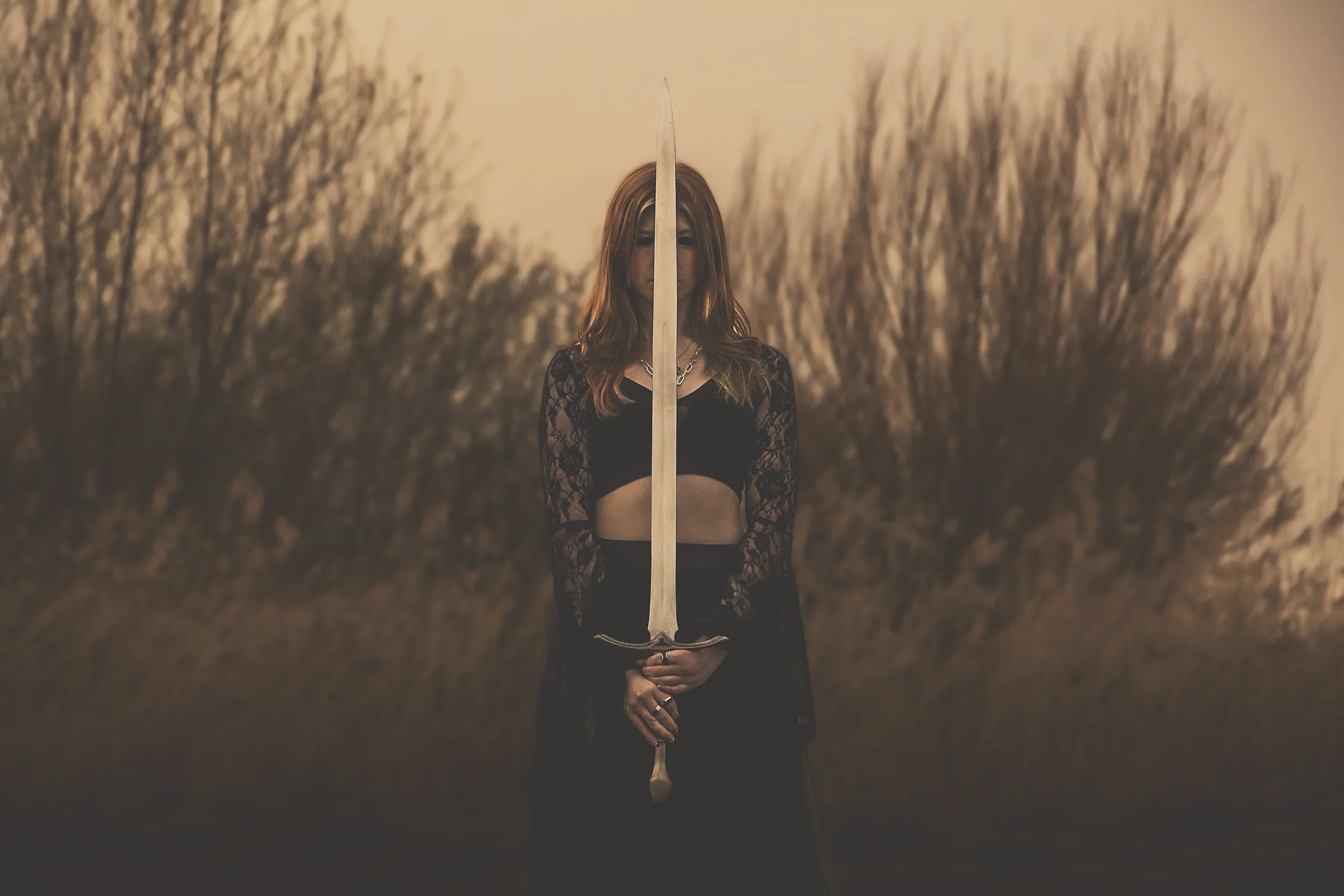 A woman with red hair holding a sword upright in front of her face, standing outdoors at sunset with tall, blurred trees in the background.