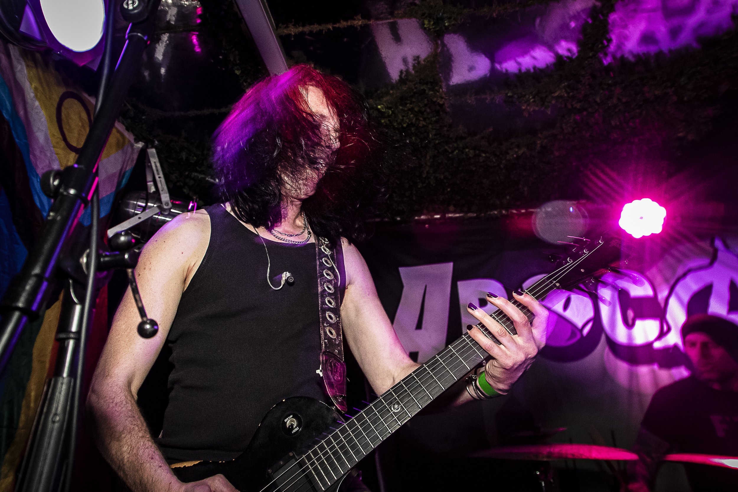 A woman playing an electric guitar on stage under purple and pink lighting, with a fellow musician in the background and a dark stage backdrop.